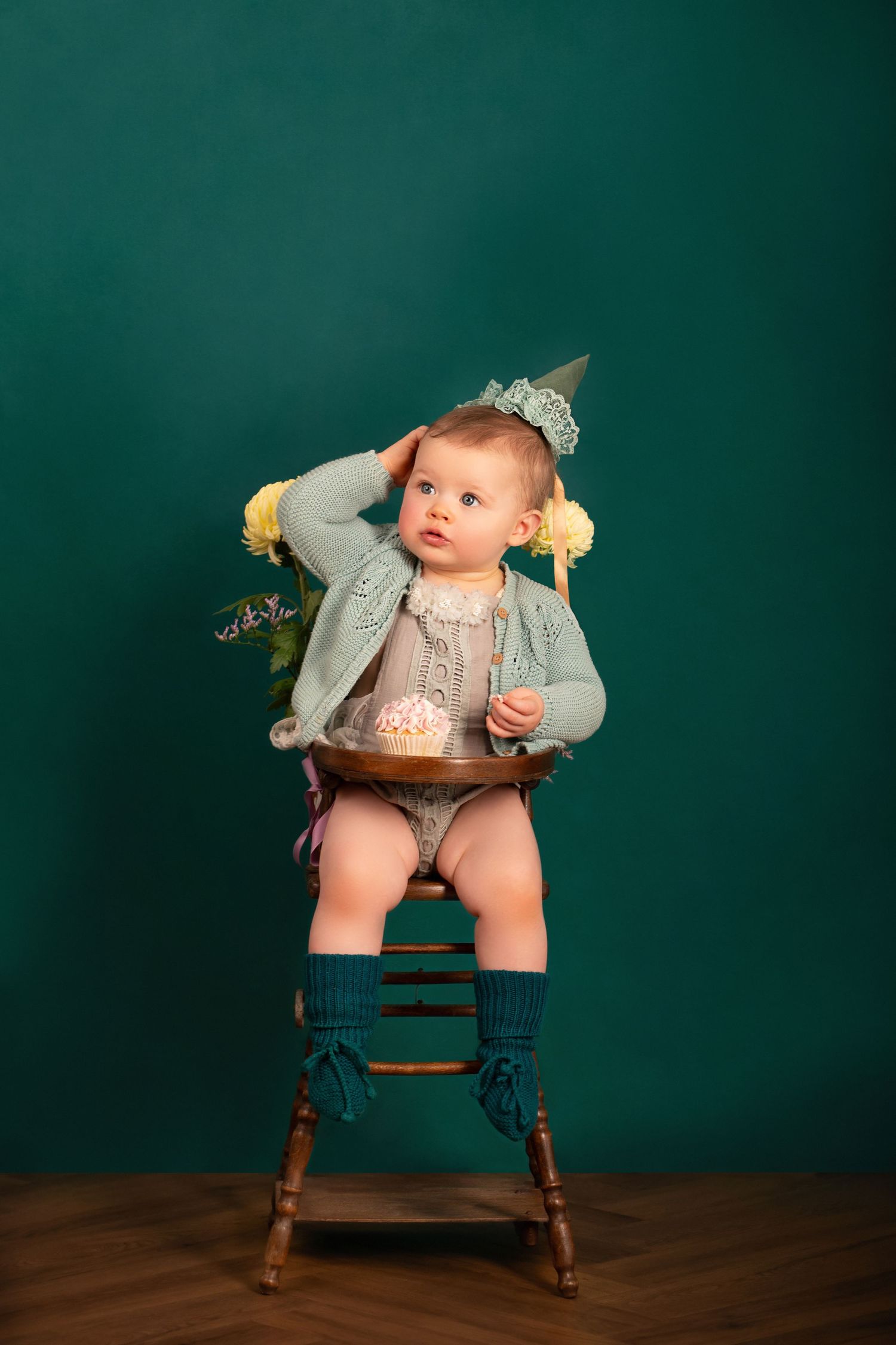 A baby wearing a gray outfit with blue socks sits in a wooden high chair against a dark green backdrop.