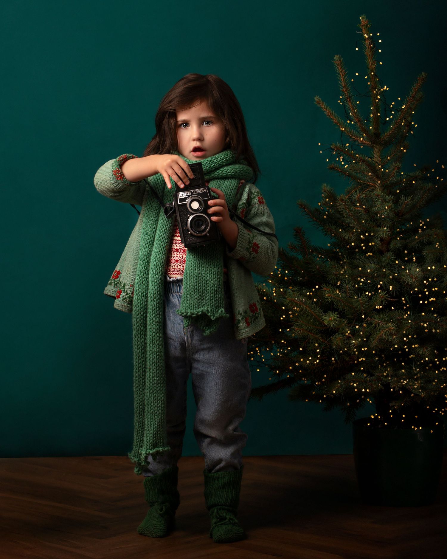 A small photographer wearing a cozy green scarf and knit socks poses with a vintage camera beside a decorated Christmas tree.