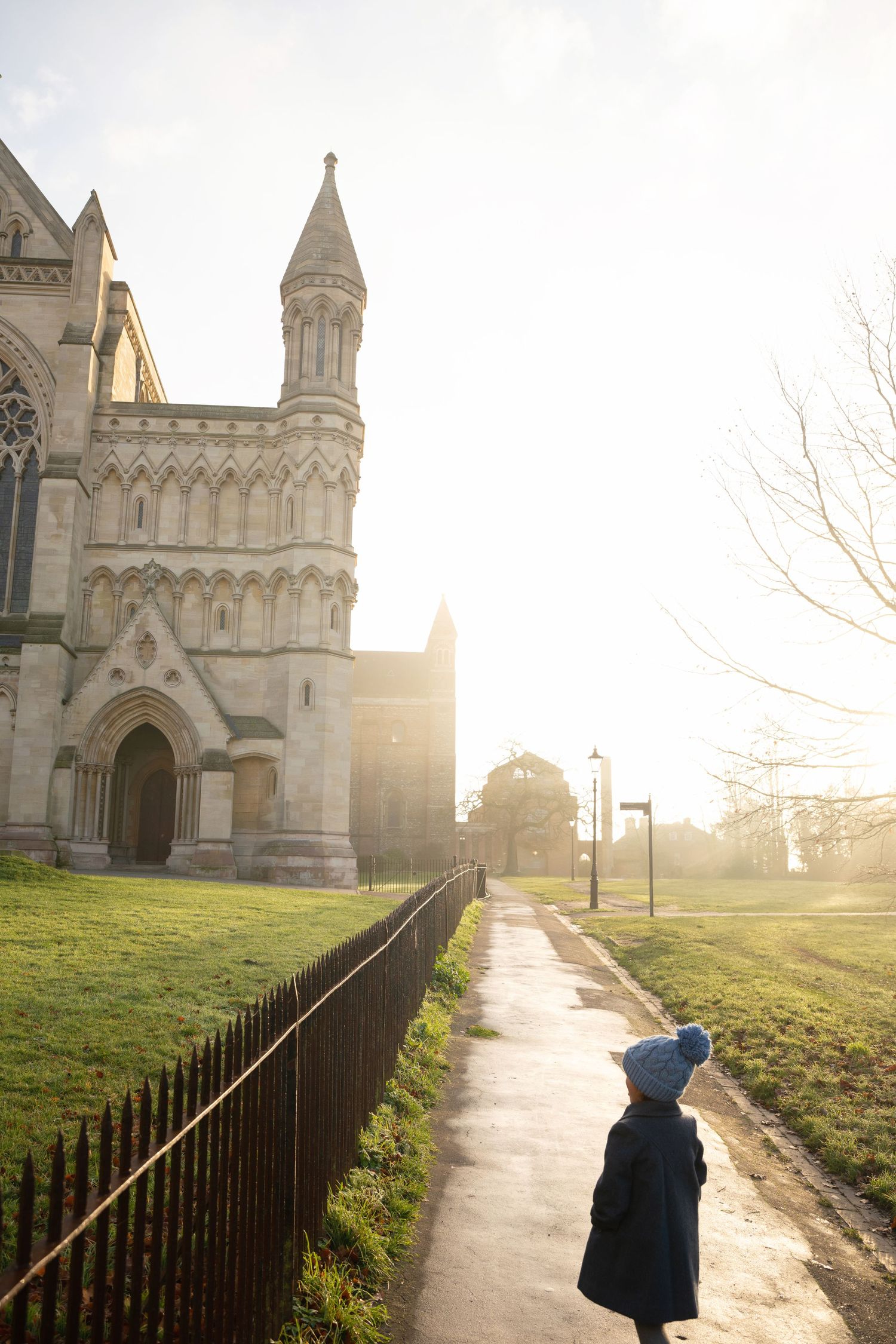 Child in blue hat walks path beside grand Gothic cathedral bathed in golden sunlight on a misty morning.