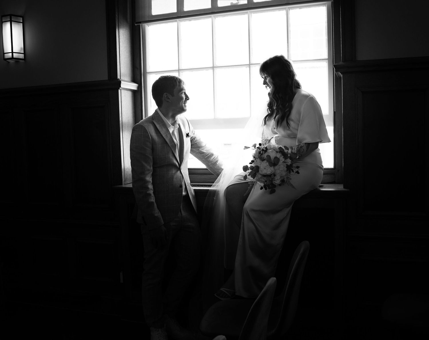 A black and white photograph of a bride and groom standing together by a window in an intimate moment. Registry in North London Enfield