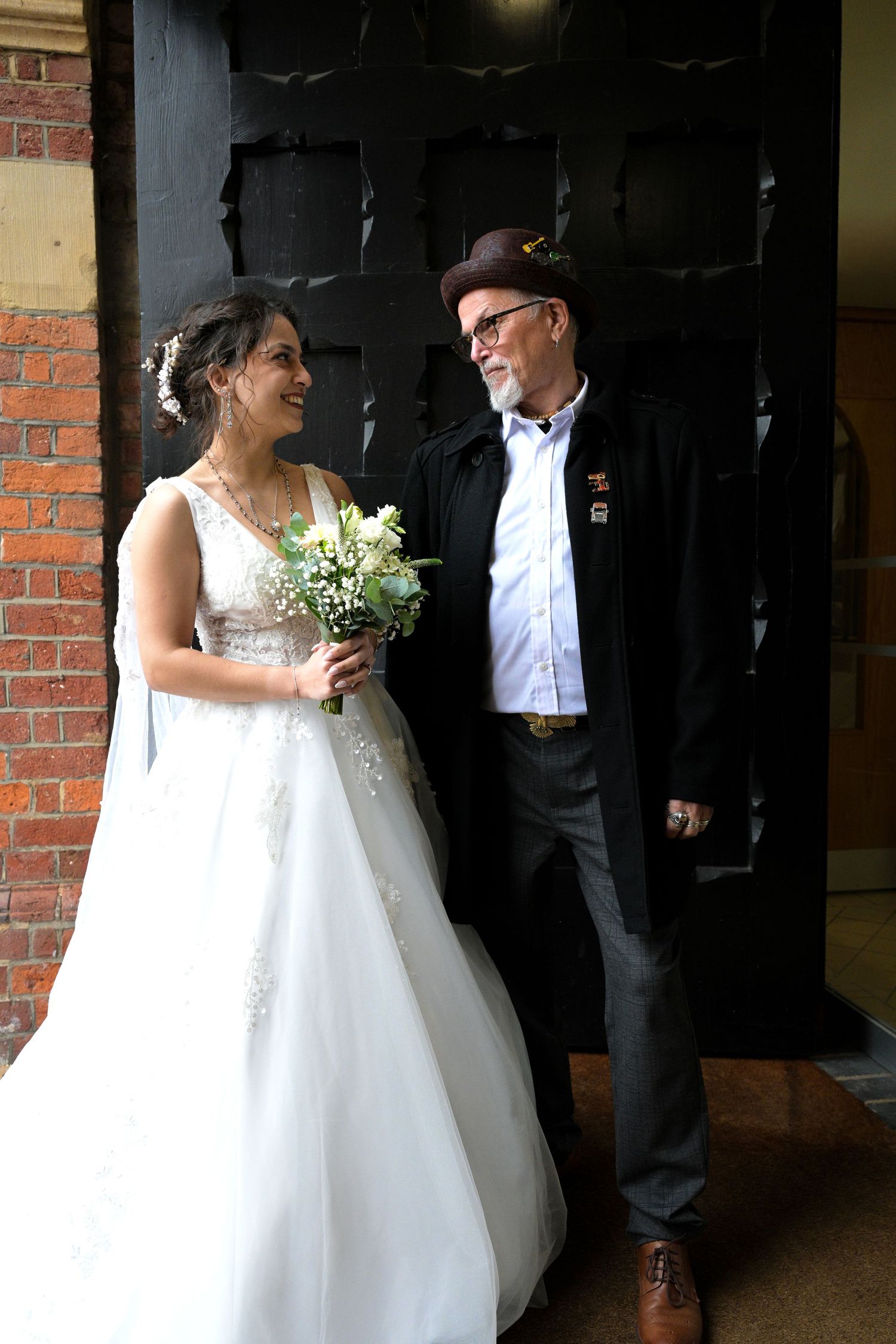 Bride in white gown holding bouquet smiles at older man in hat and dark coat outside brick building.
