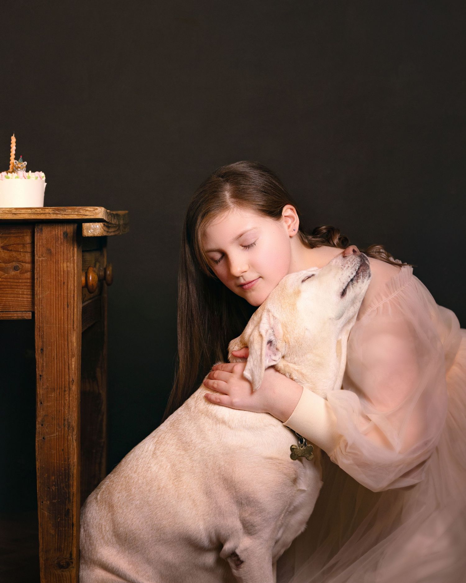 A young girl in a pink dress hugs a yellow Labrador beside a birthday cake on a wooden table.
