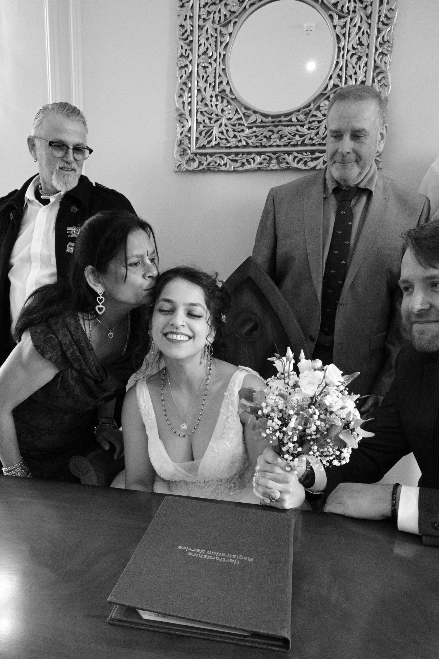 Black and white wedding photo of smiling bride holding bouquet, surrounded by family members at signing table.