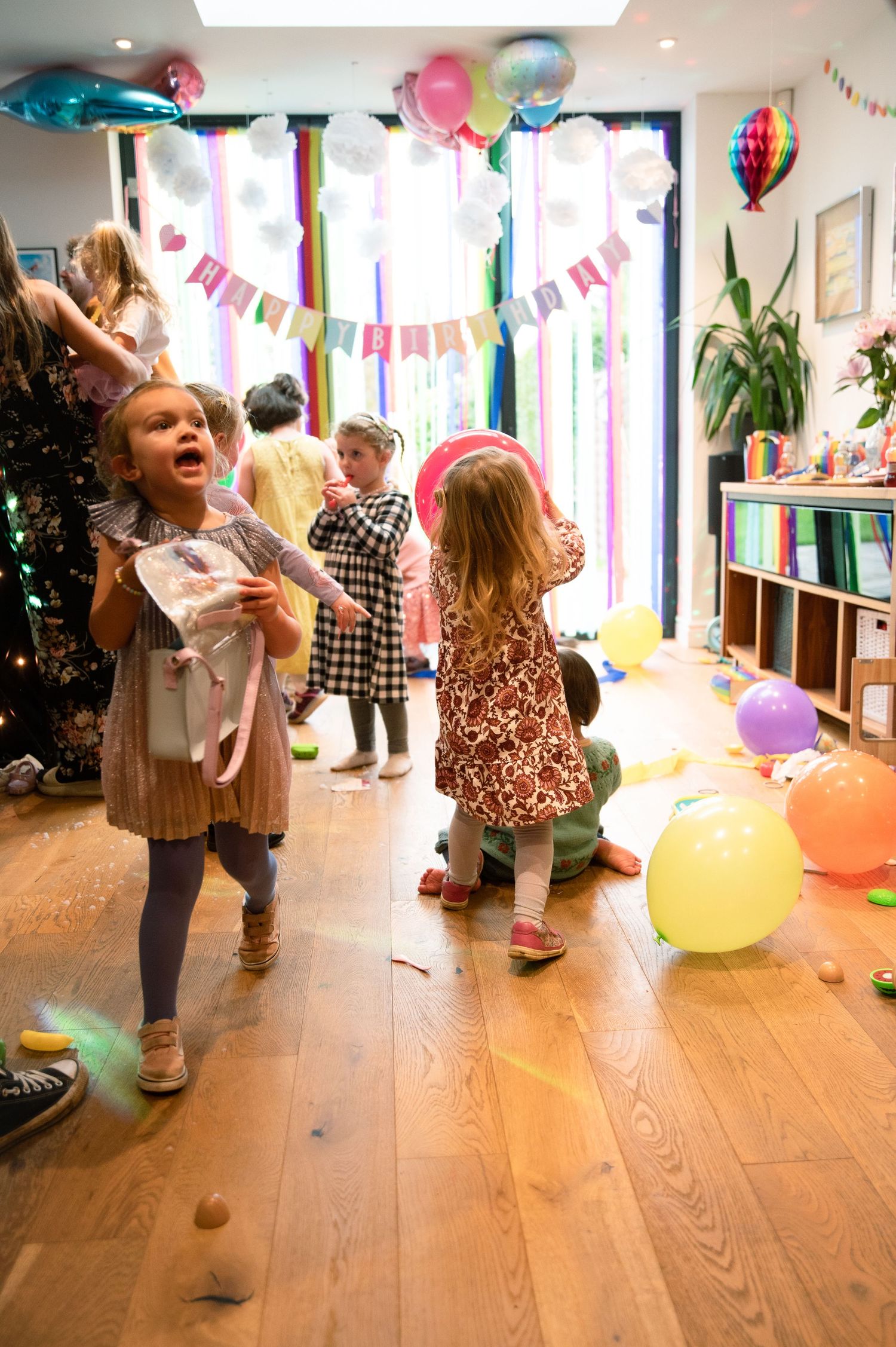 Children dancing and playing at a birthday party with colorful balloons and streamers decorating a bright indoor space.