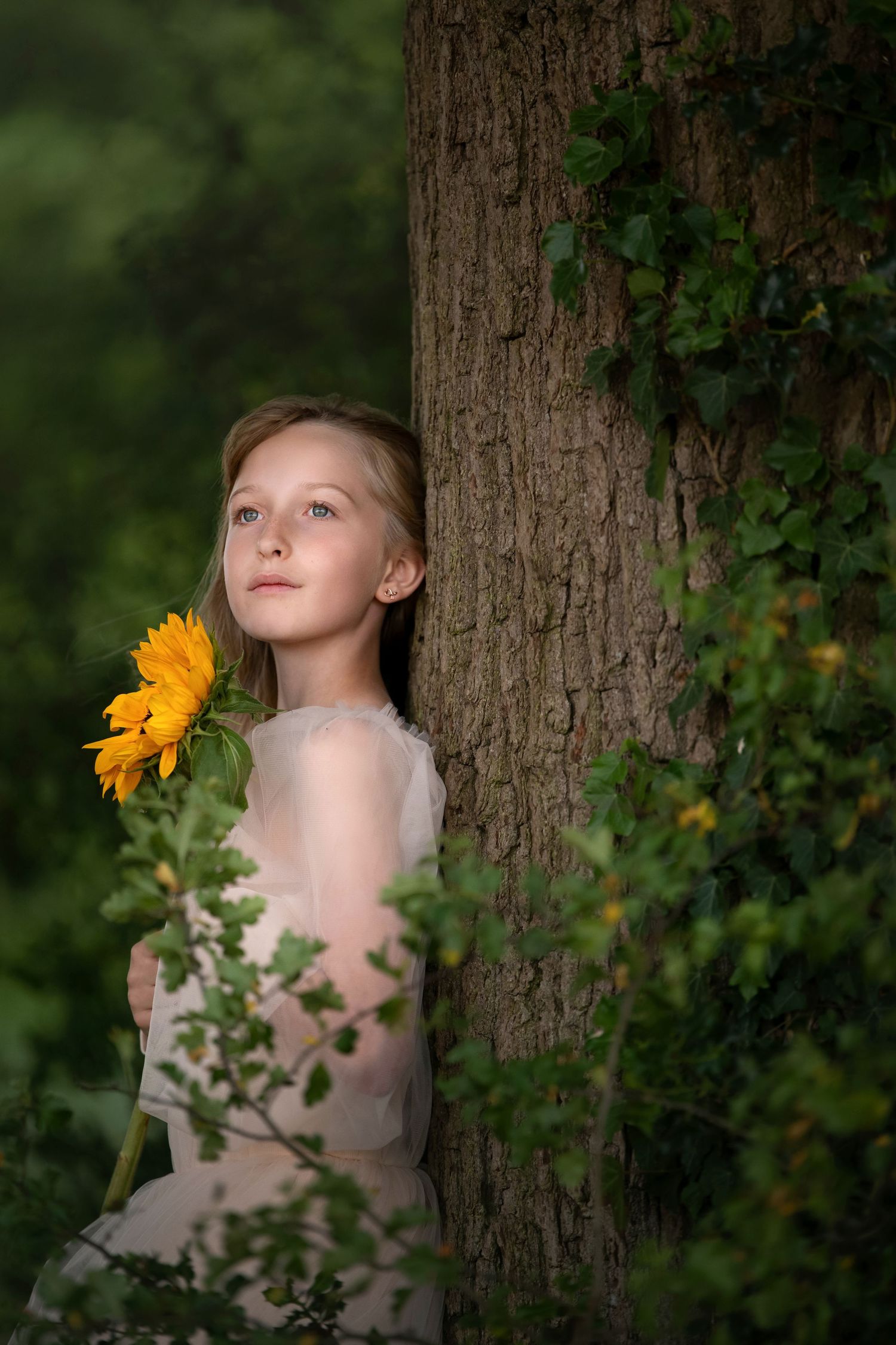 Young girl holding a sunflower leaning against a tree in a lush green forest wearing a white dress.