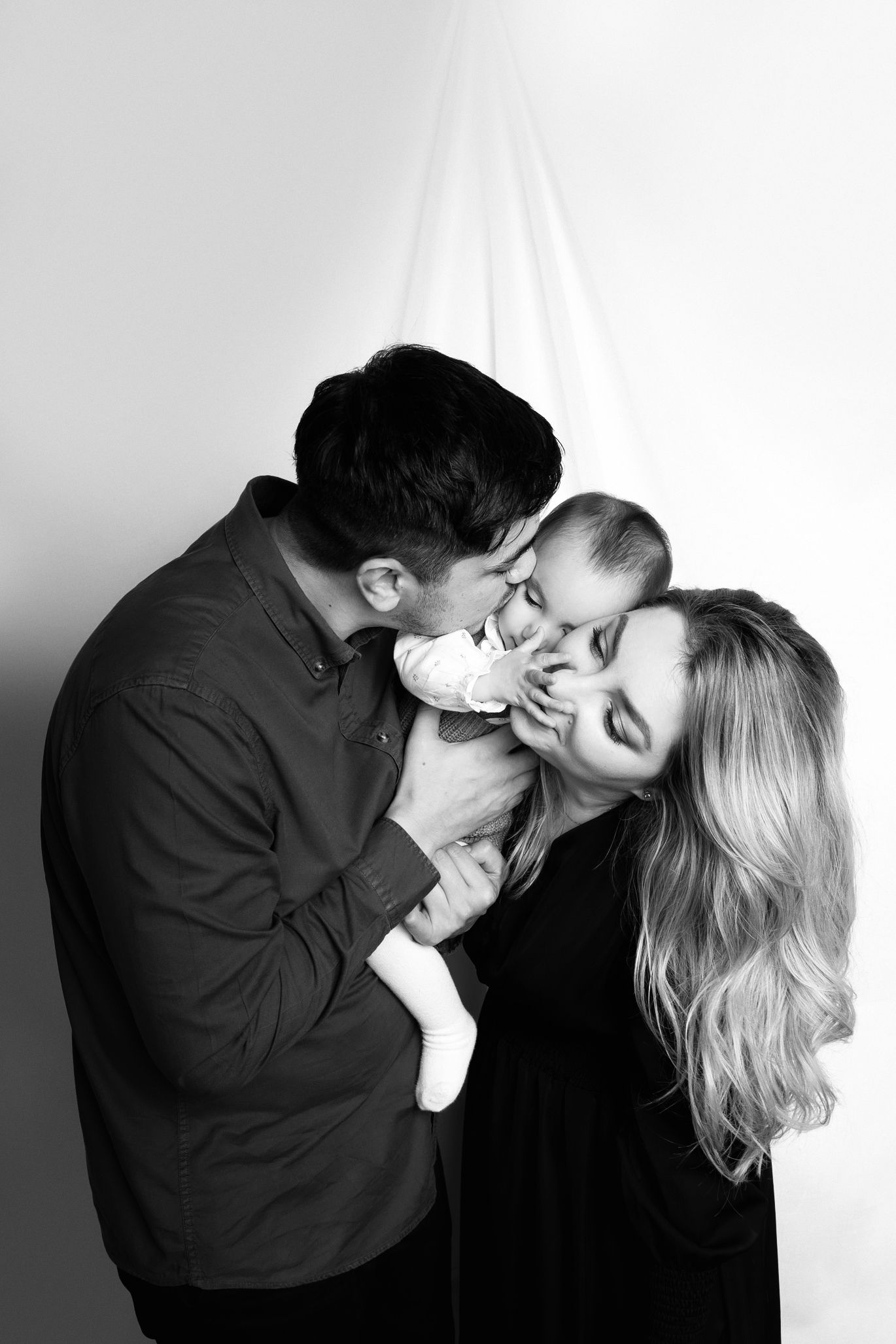 Black and white photo of a couple kissing their newborn baby in a studio portrait setting.