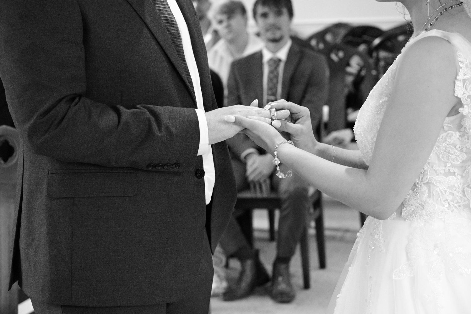 Black and white photo of groom placing wedding ring on brides finger during ceremony with guests seated in background.