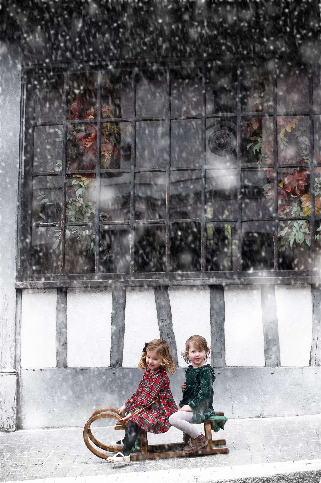 Two young girls sitting on a vintage sled in falling snow outside a rustic building with large windows.