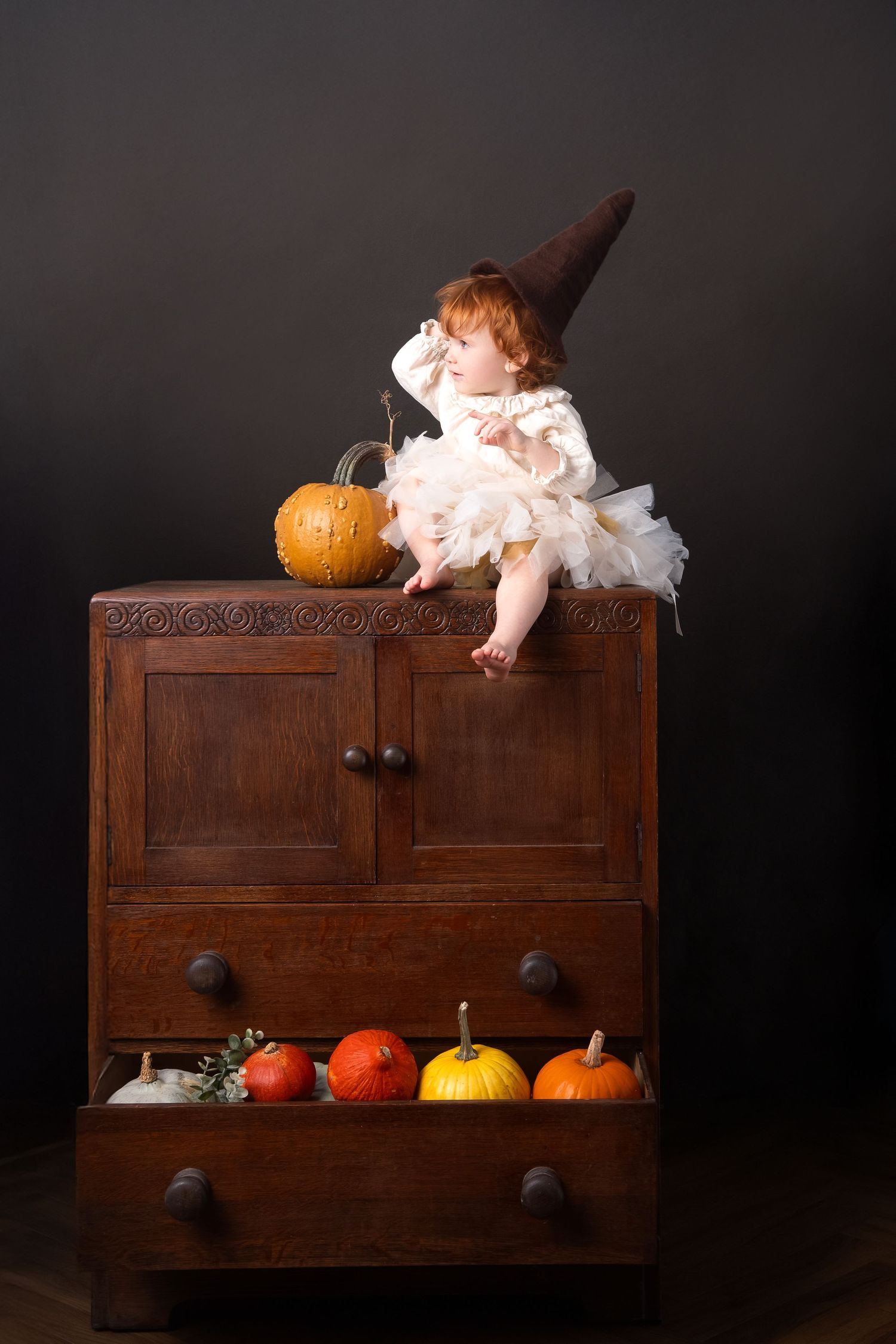 Baby in witch hat and white dress sits atop wooden dresser with pumpkins for Halloween.
