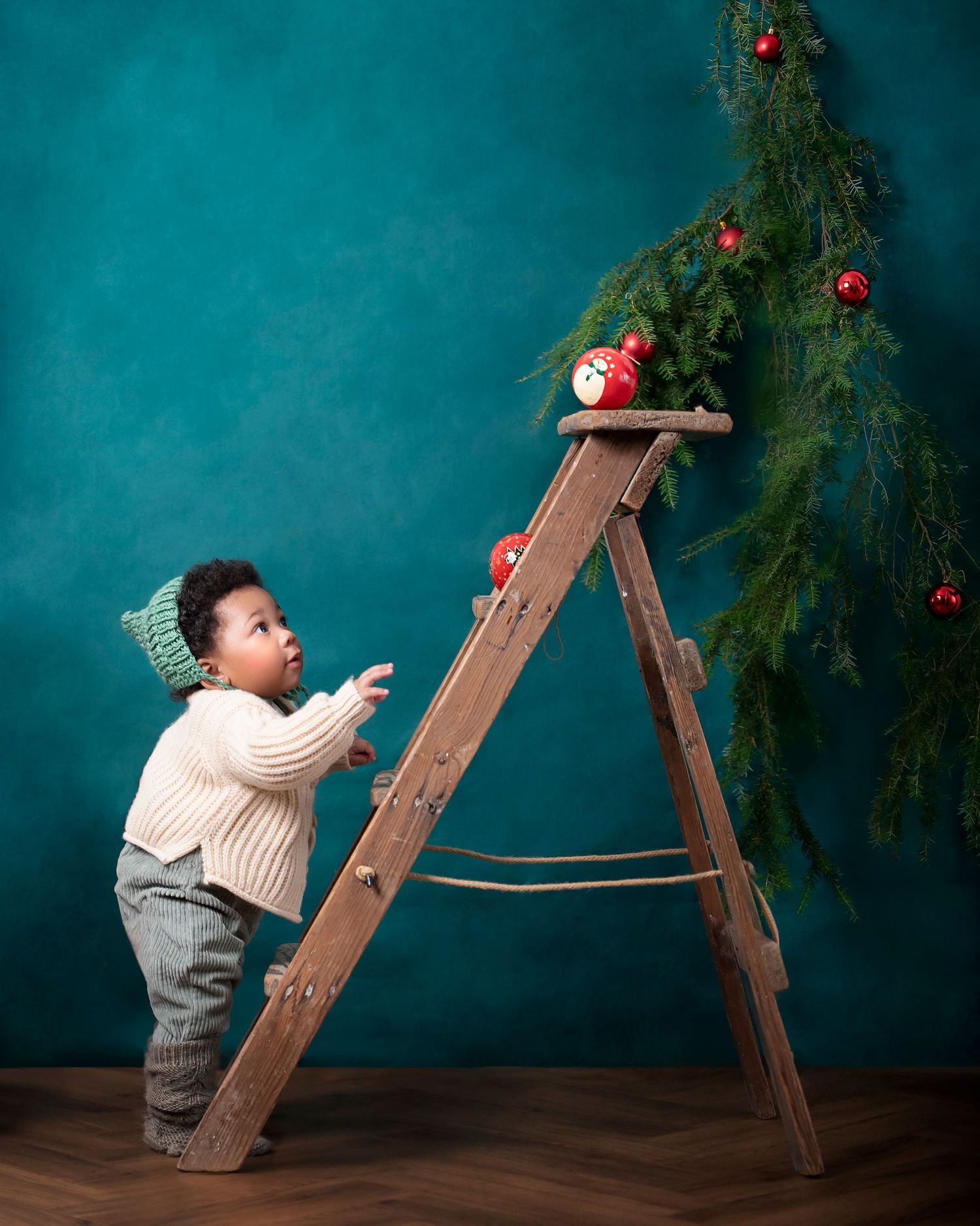 Small child in vintage outfit explores a wooden ladder near a decorated Christmas tree against a teal backdrop.