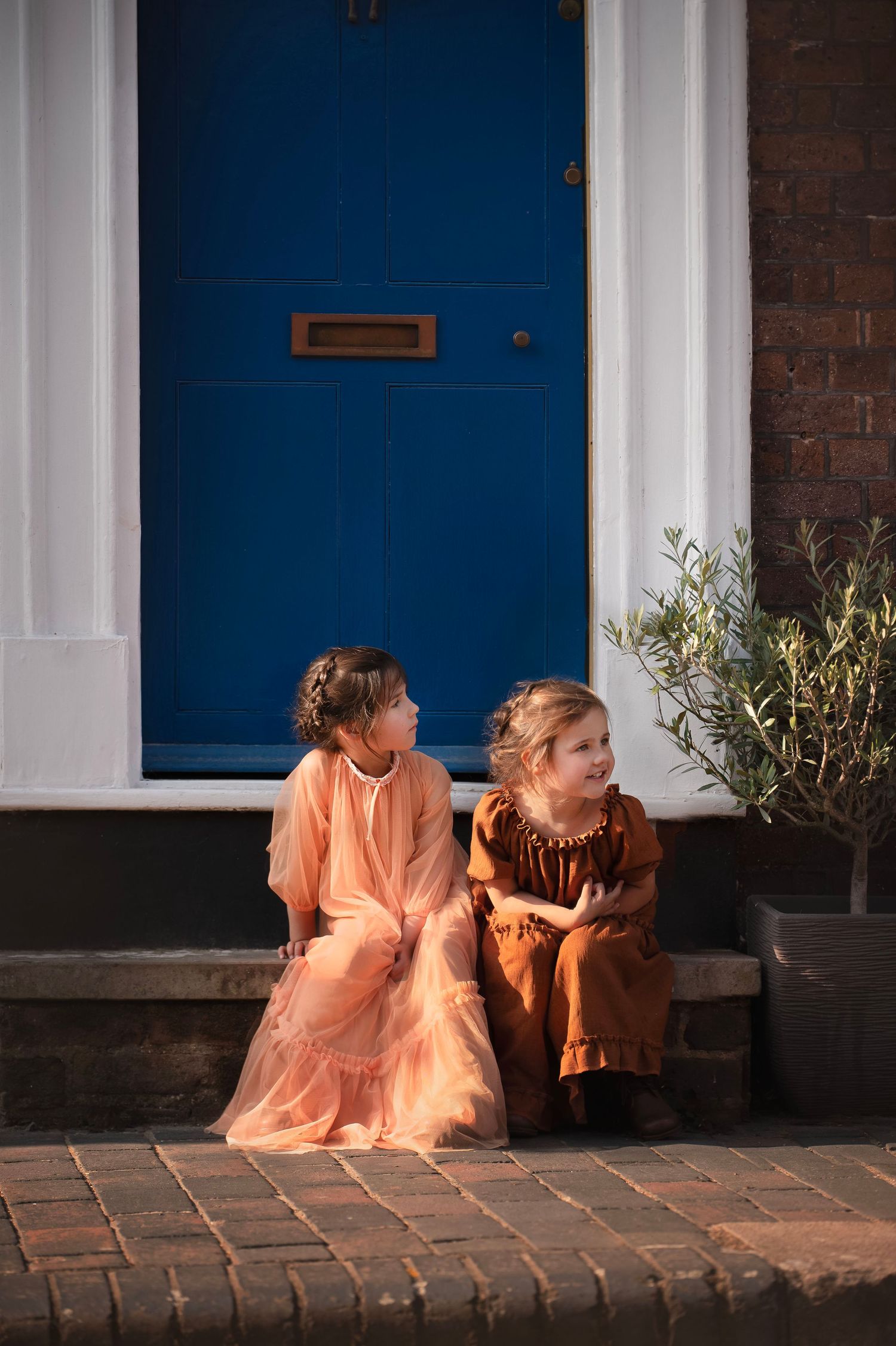Two figures in peach-colored outfits sit together on a brick porch with white columns and blue doors.