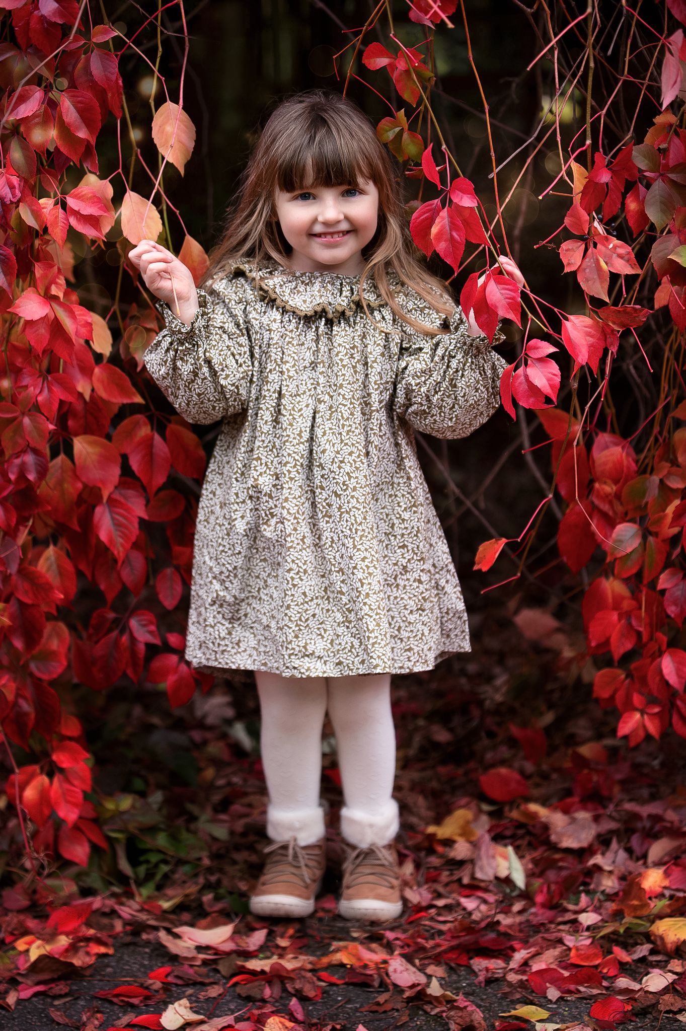 A smiling young girl in a floral dress and white tights stands among vibrant red autumn leaves in a garden.