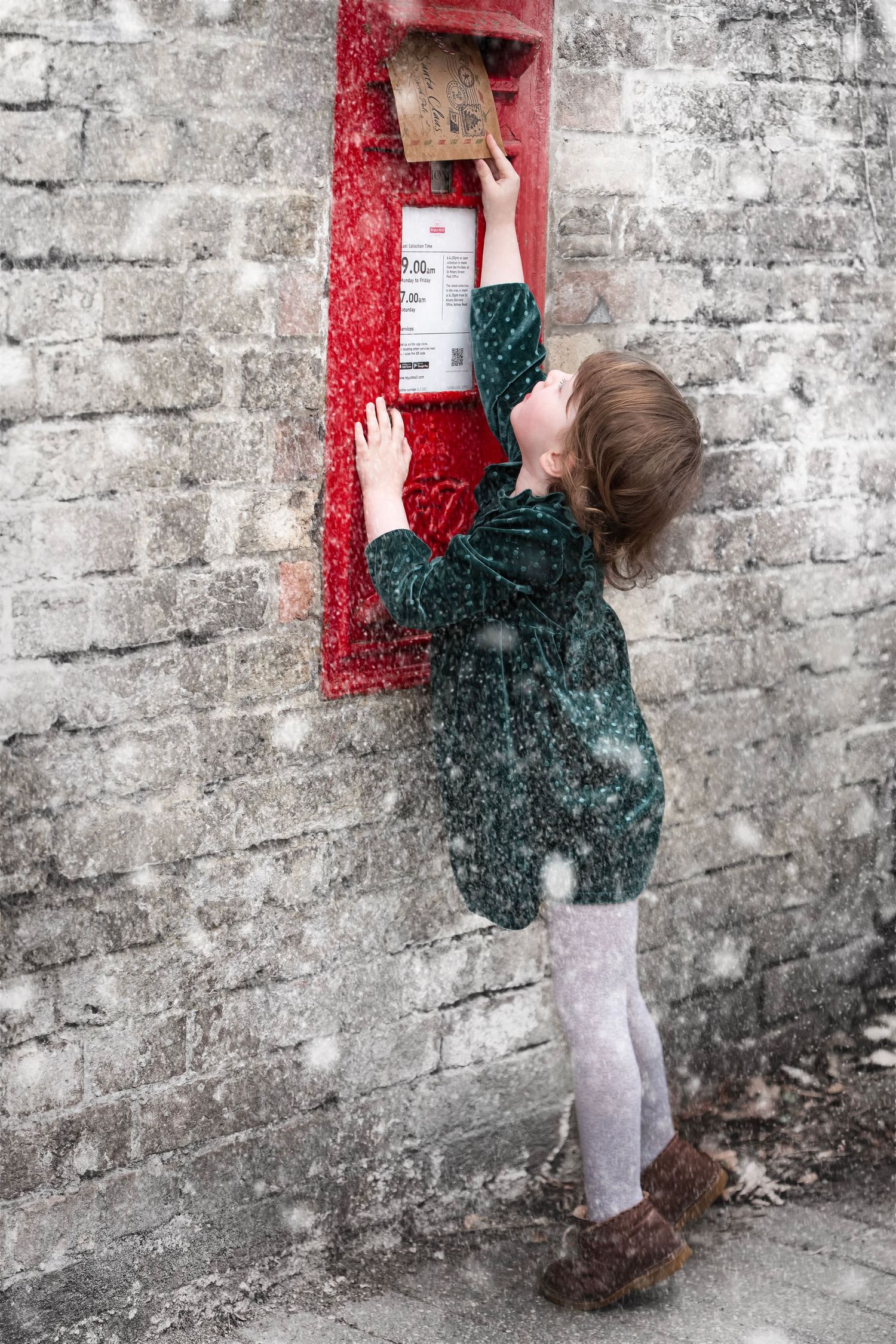 Young girl posting a letter in a red British postbox during a snowy winter day, wearing a green coat and tights.