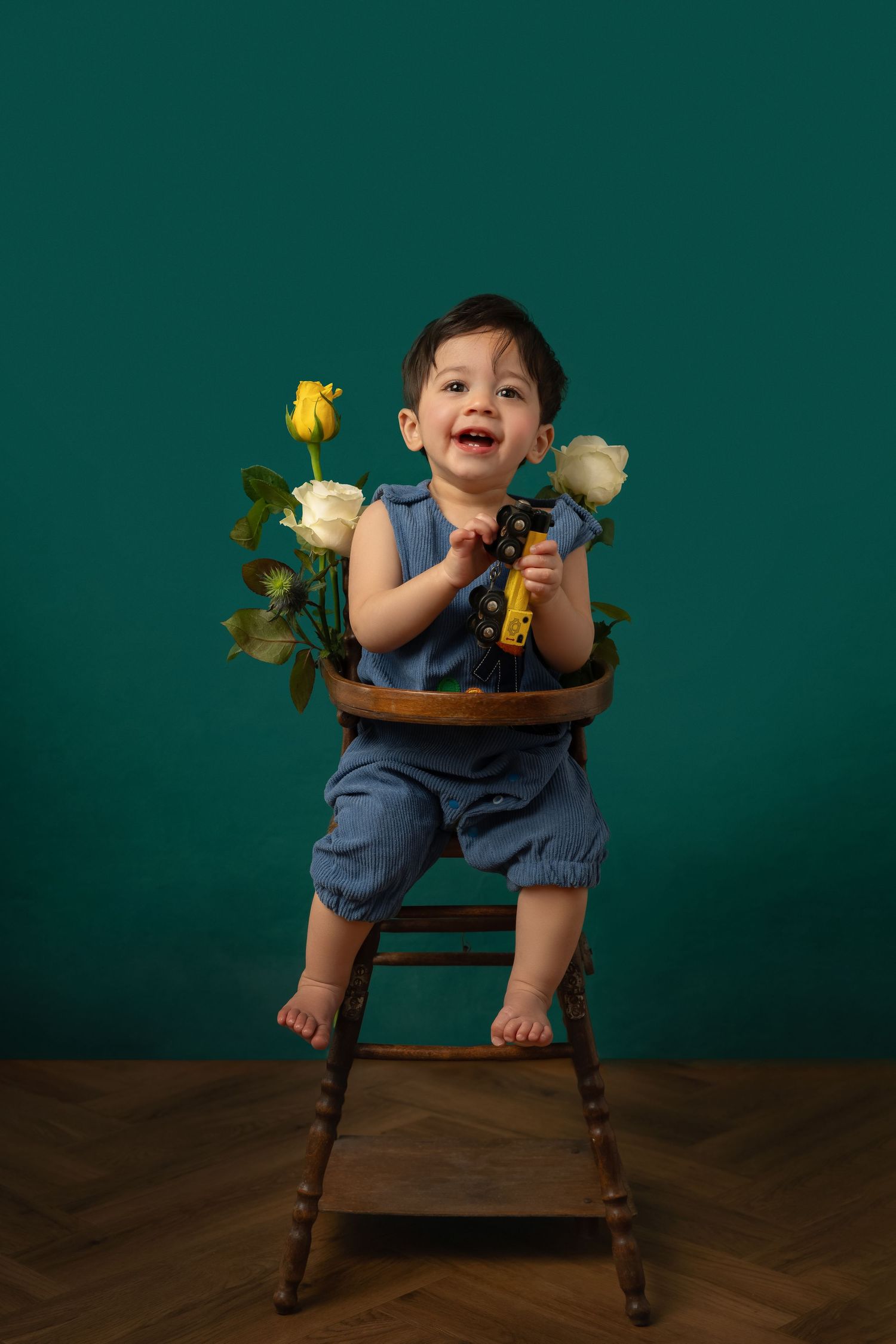 Happy toddler in blue romper sitting on wooden chair with yellow and white roses against teal background.