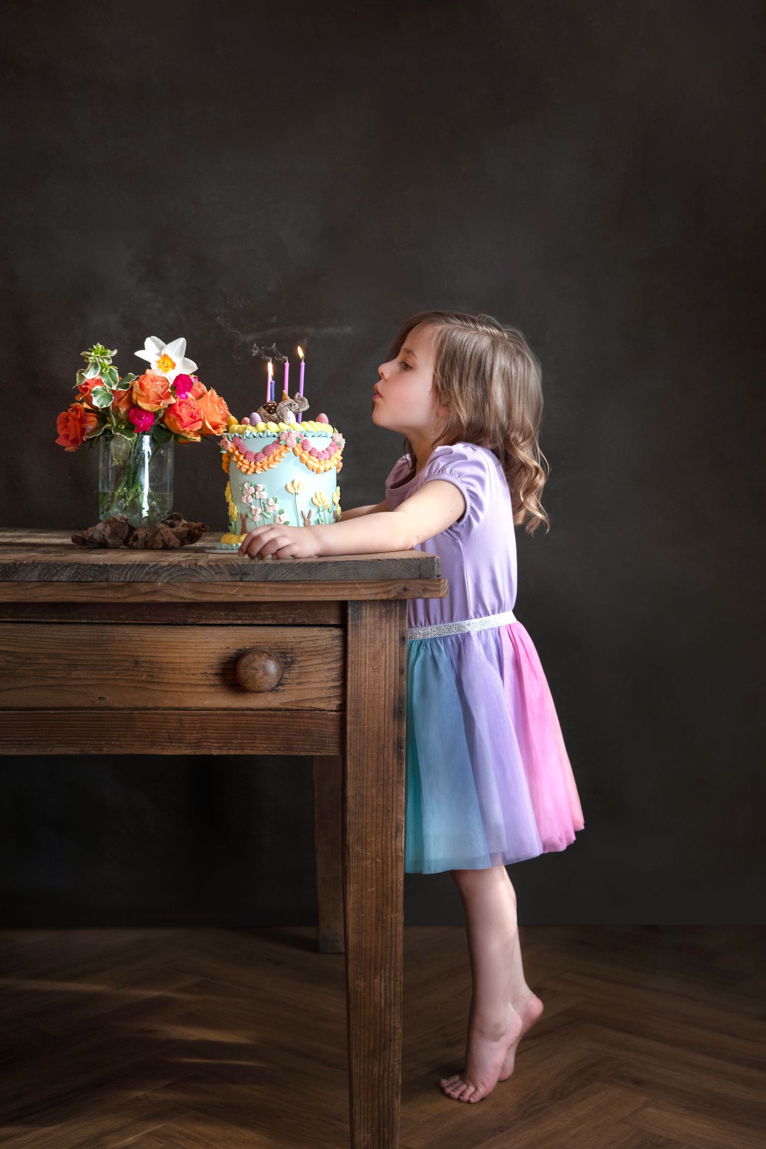 A barefoot girl in a colorful dress blows out candles on a birthday cake at a wooden table.