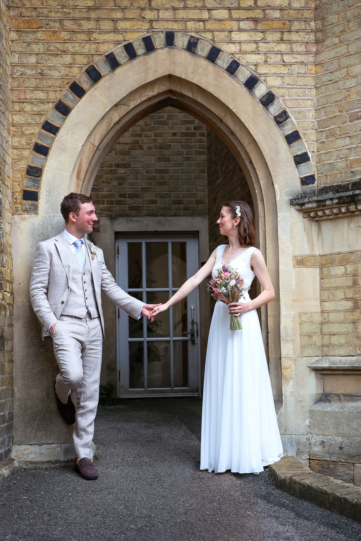 A couple in wedding attire poses playfully in front of a historic stone archway with brick details.