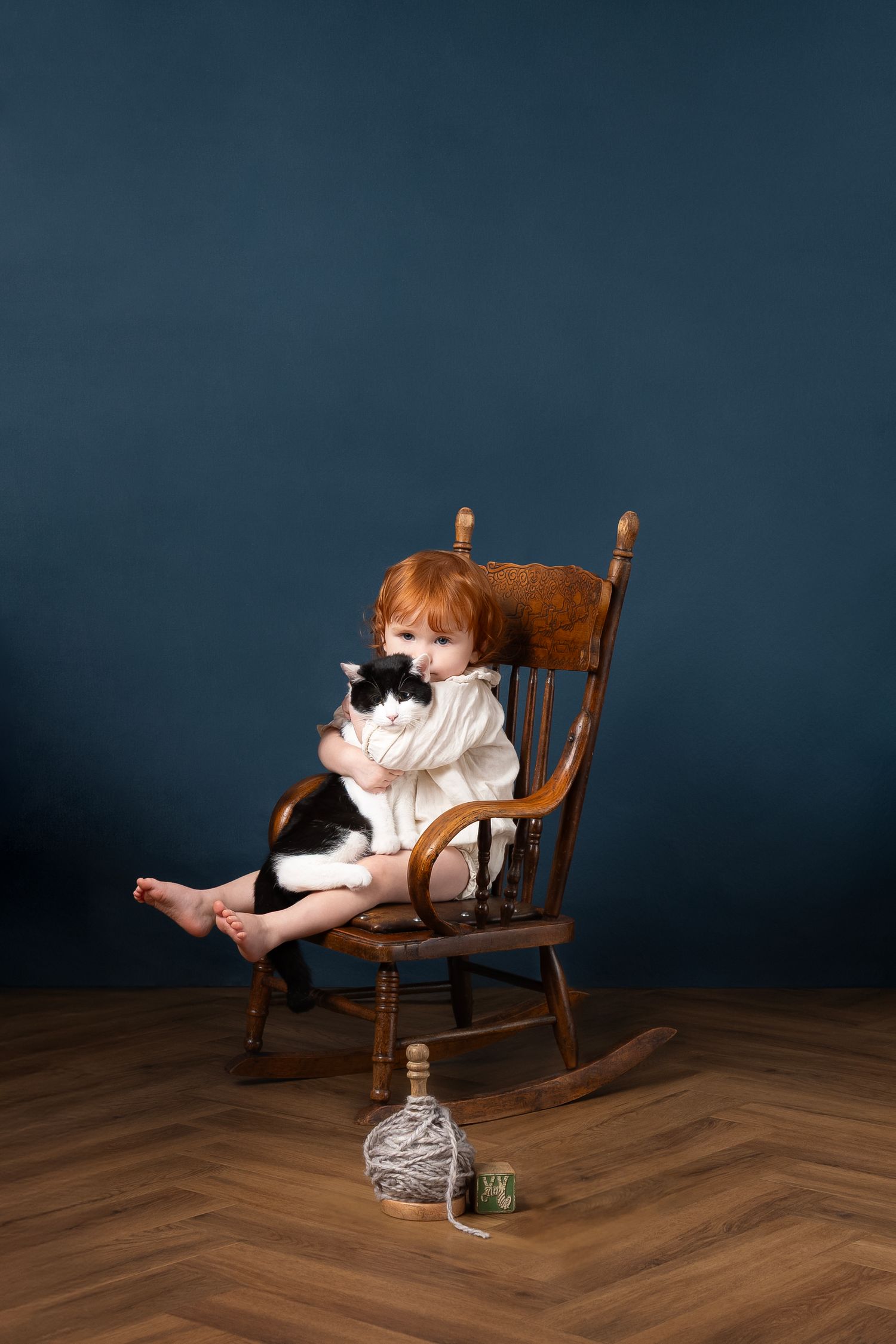 A small child in a white dress sits in a wooden rocking chair holding a kitten on a hardwood floor against a dark backdrop.