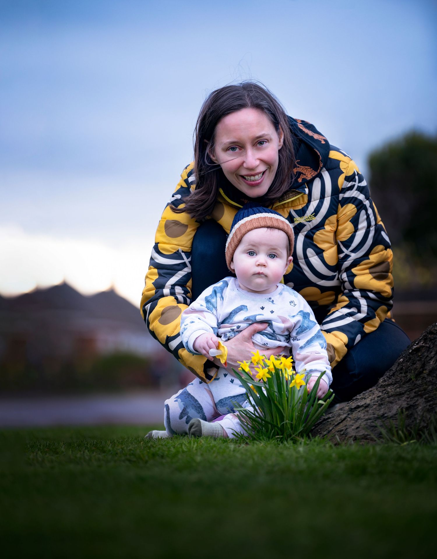 Person in yellow patterned jacket kneeling next to baby outdoors with daffodils blooming in the grass.