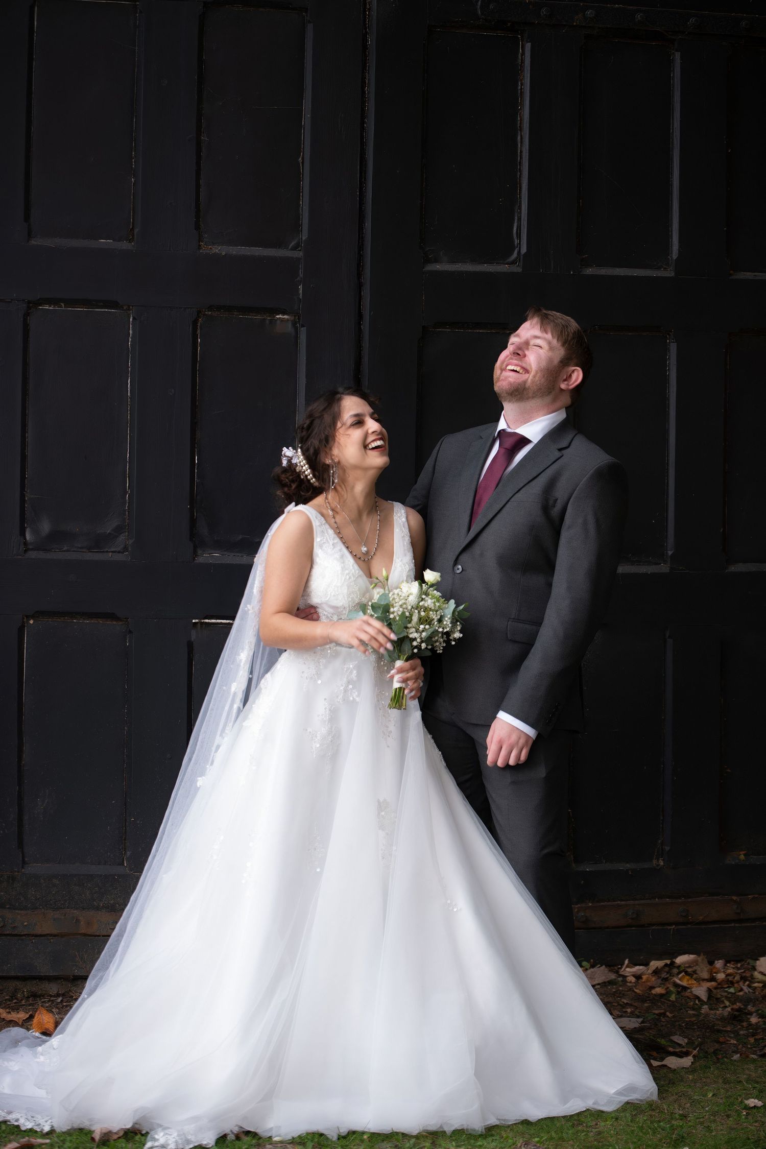 A joyful bride in a white gown holding a bouquet and groom in a gray suit laughing together before dark wooden doors.