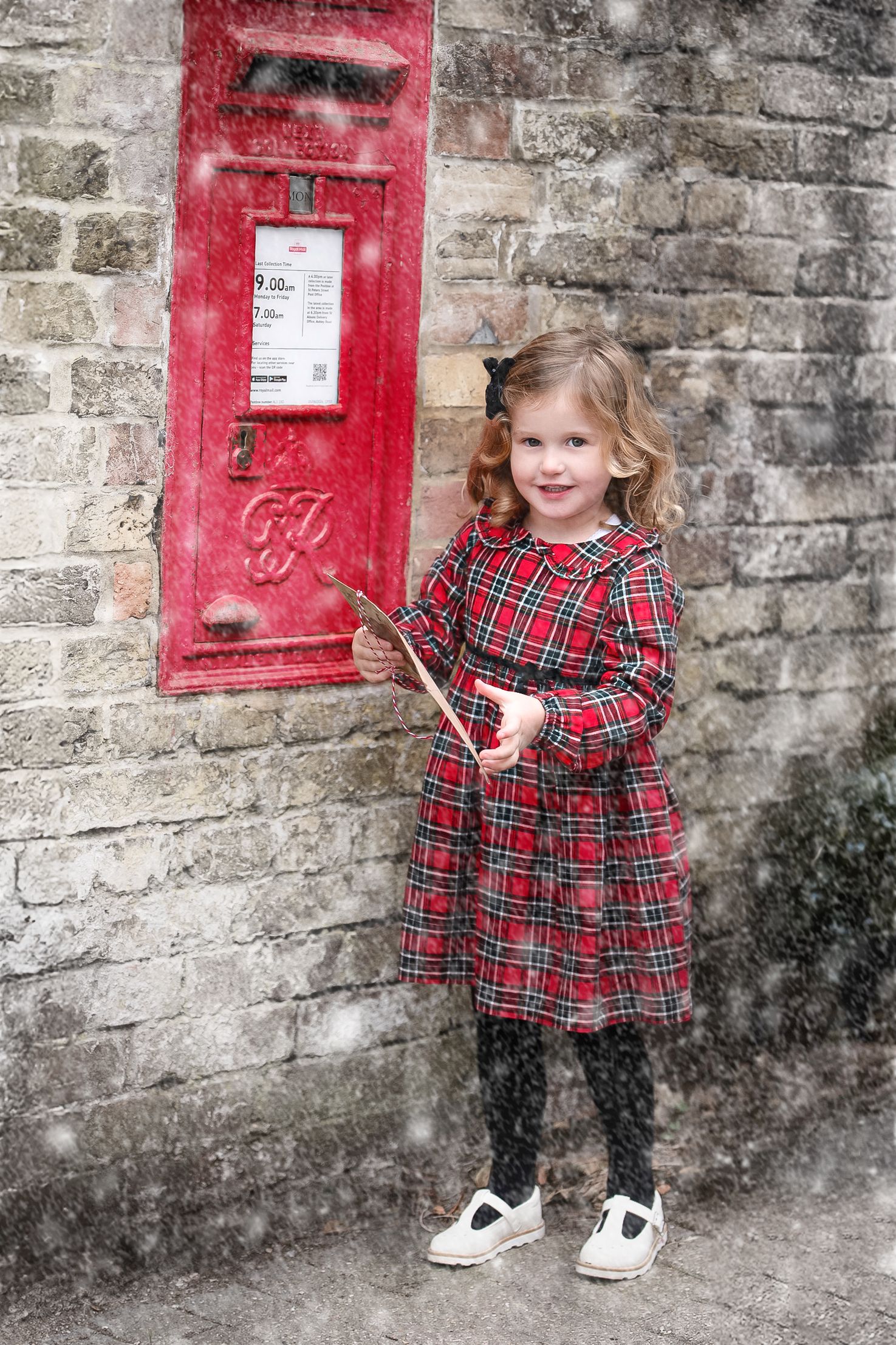 A young girl in a red tartan dress holds a letter beside a red British post box in the snow.