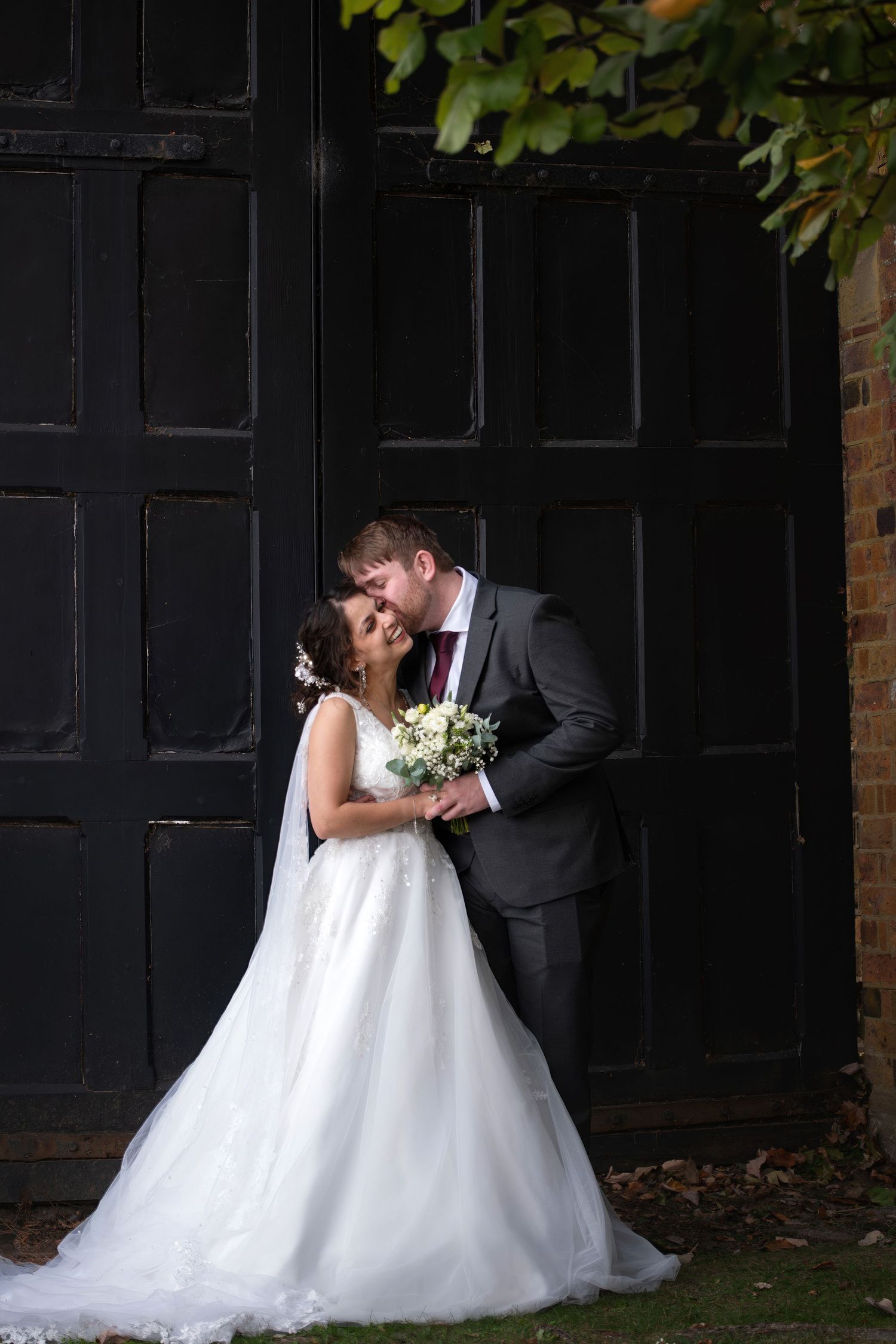 Bride and groom share a romantic moment in front of a black door, bride holding a white bouquet in a flowing gown.