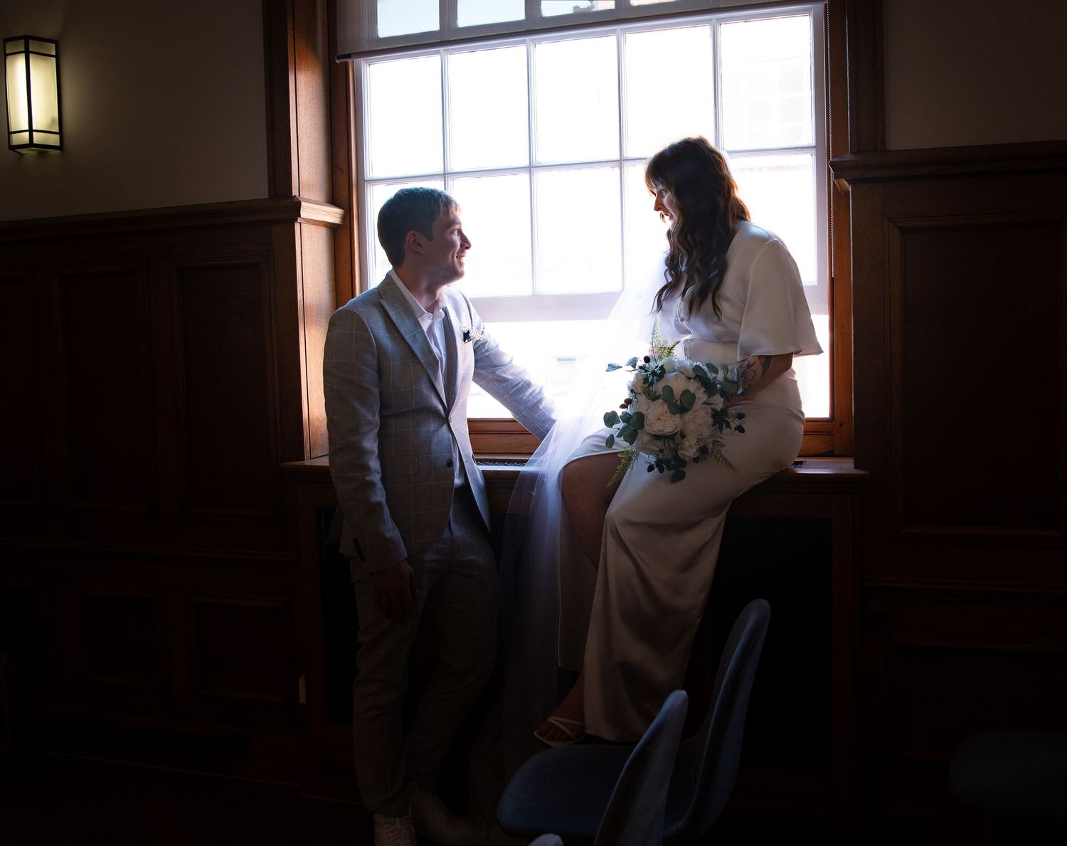 A couple shares an intimate moment in a dimly lit room with window light creating a romantic silhouette.