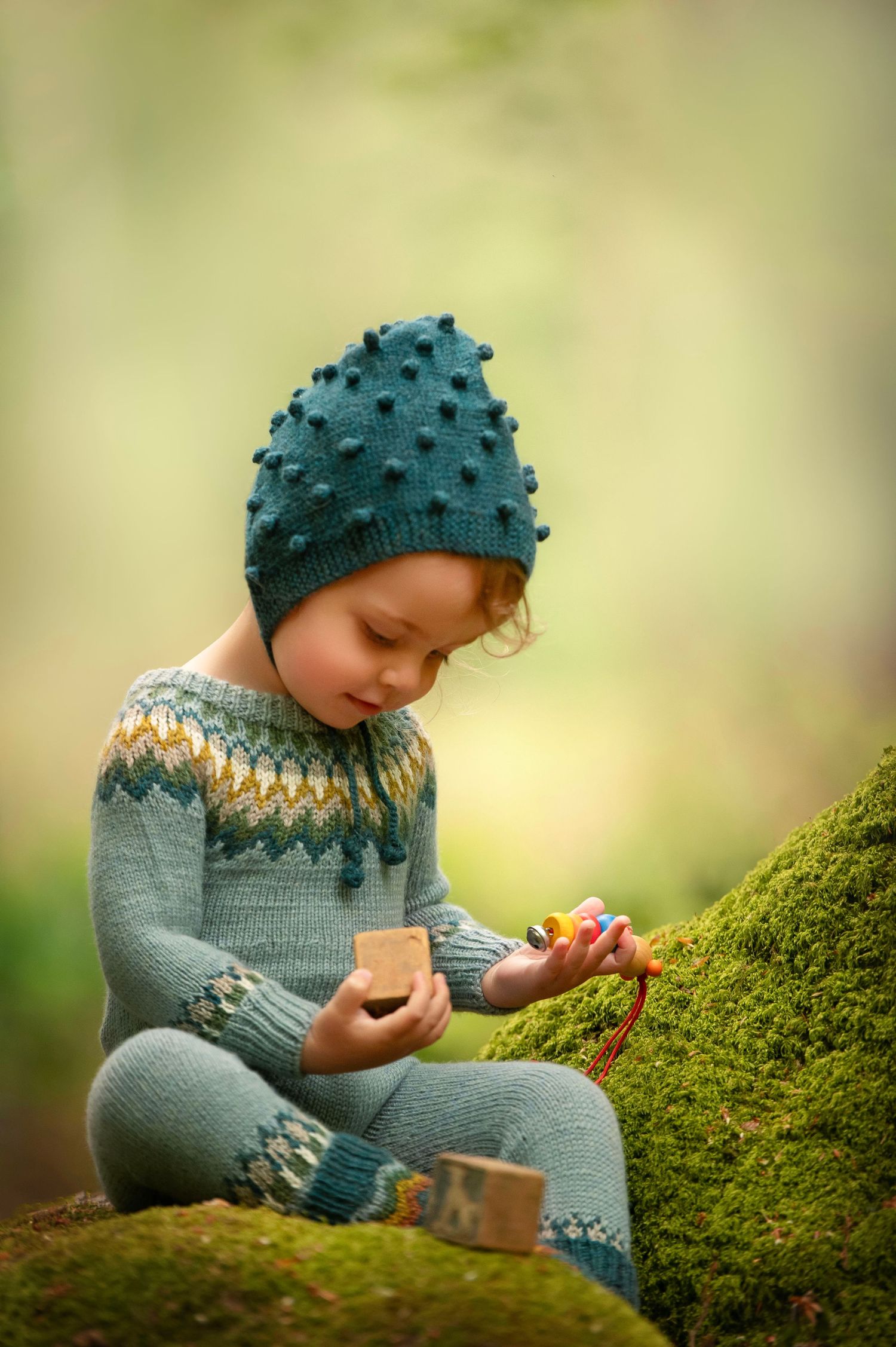 A child wearing a teal knit hat explores moss-covered ground in soft natural light.