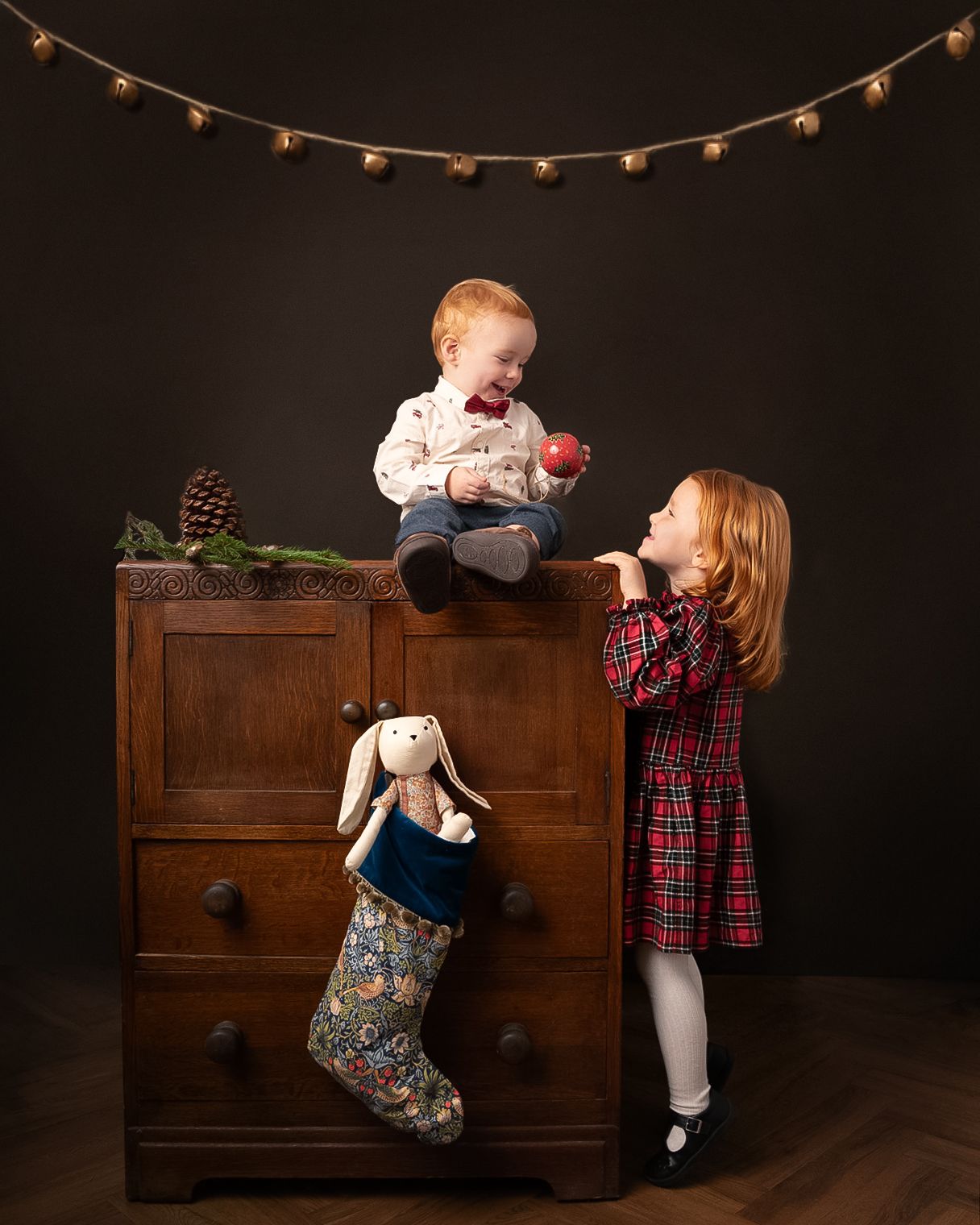 Two young children climb on a vintage wooden dresser during a playful holiday photo session with string lights above.