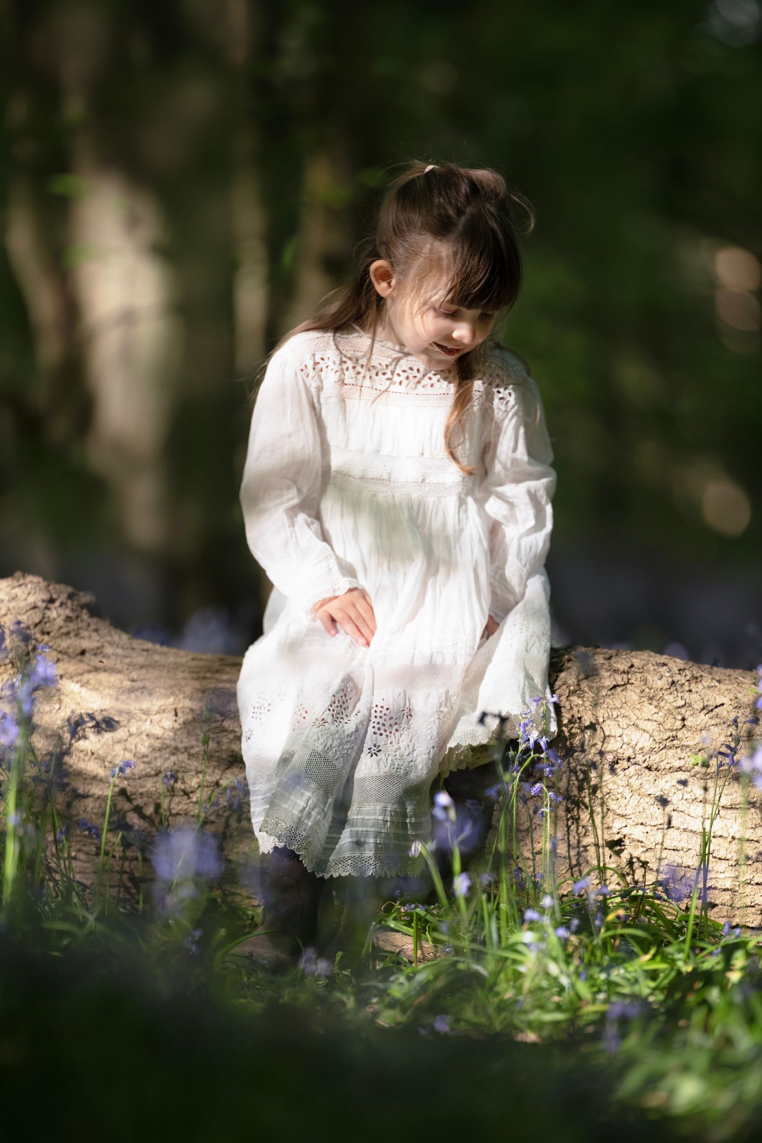 Young girl in white lace dress sitting on a log surrounded by bluebells in a sunlit woodland setting.