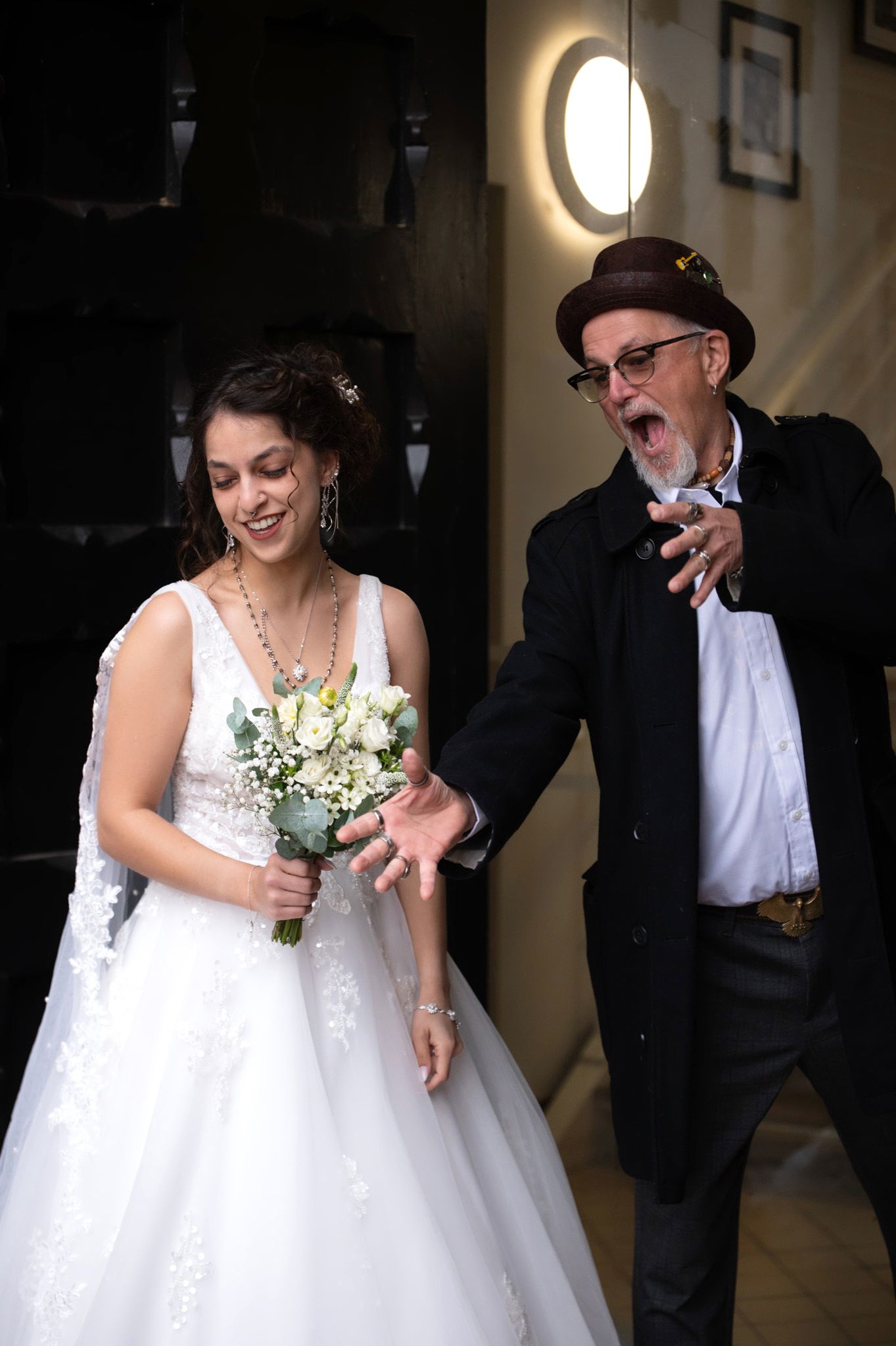 Bride in white gown holding bouquet laughs beside older man in black coat and brown hat at wedding venue.