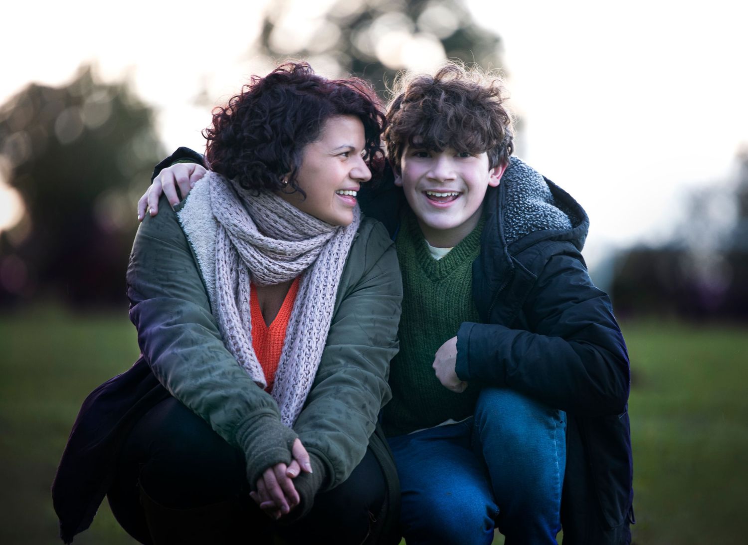 Two people in winter coats share a close moment while sitting together outdoors at dusk.