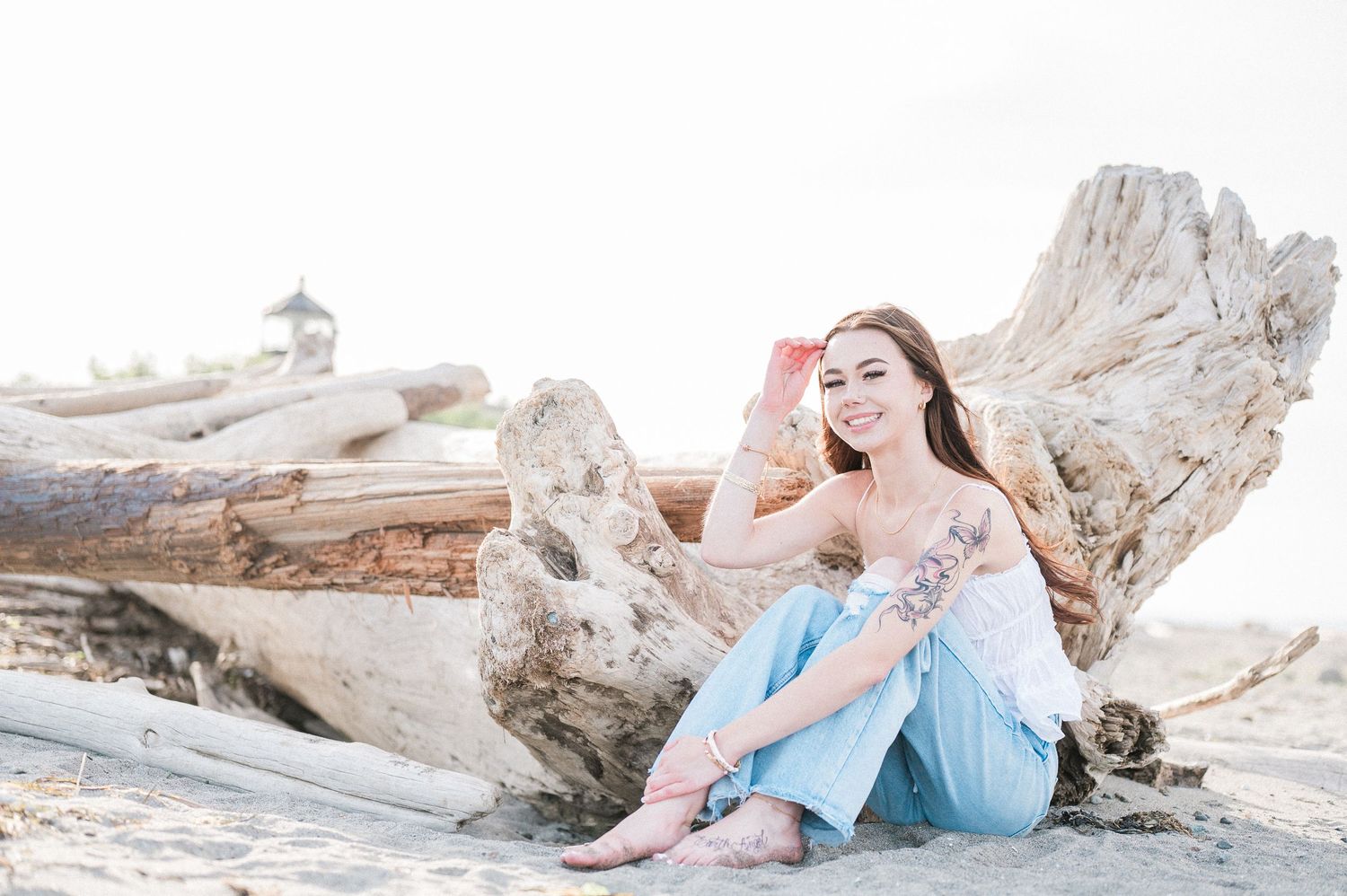 A high school senior girl in a flowy top sits gracefully on white driftwood at the beach during sunset.