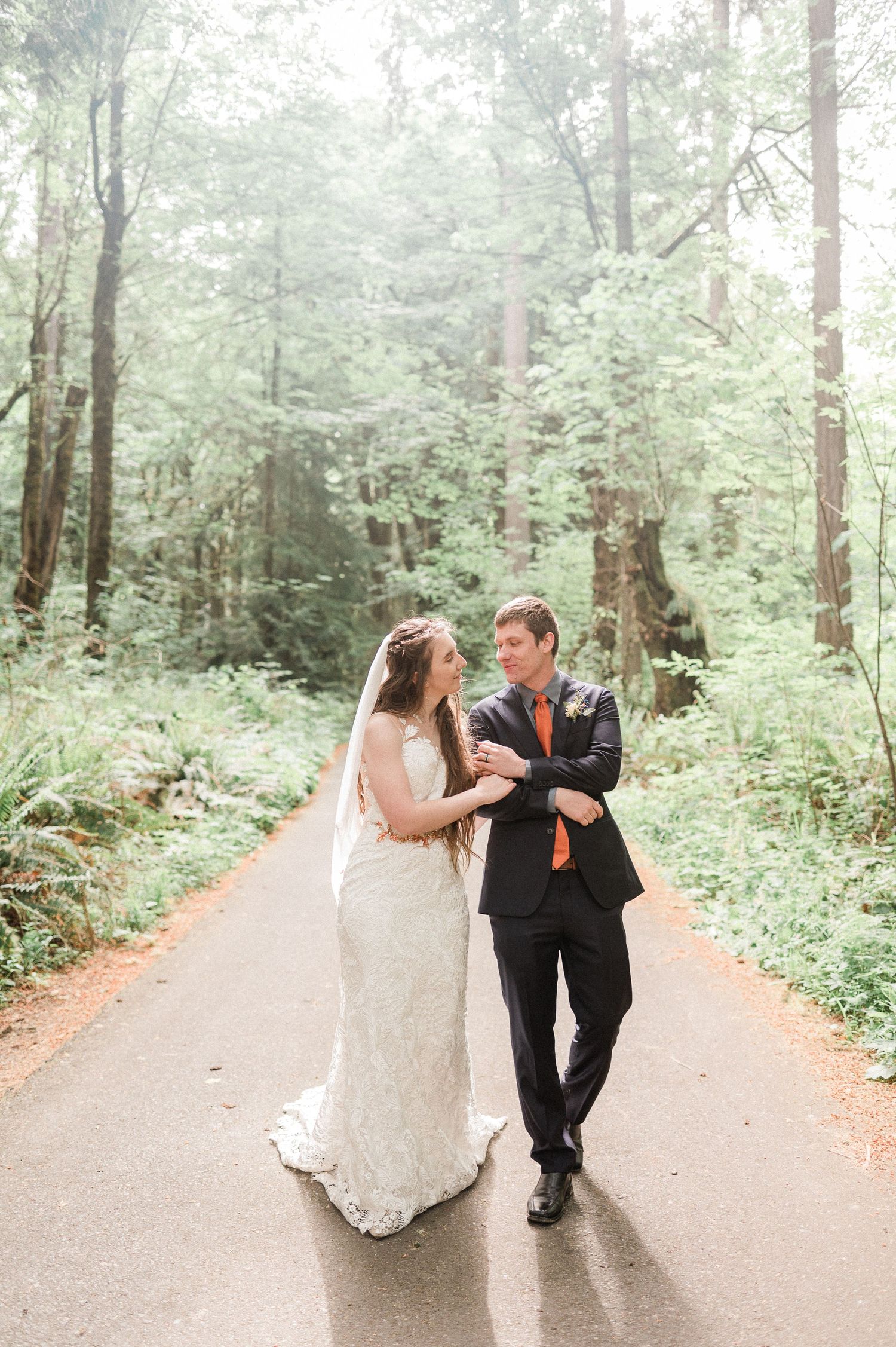 A couple in wedding attire walks along a misty forest path surrounded by tall evergreen trees.