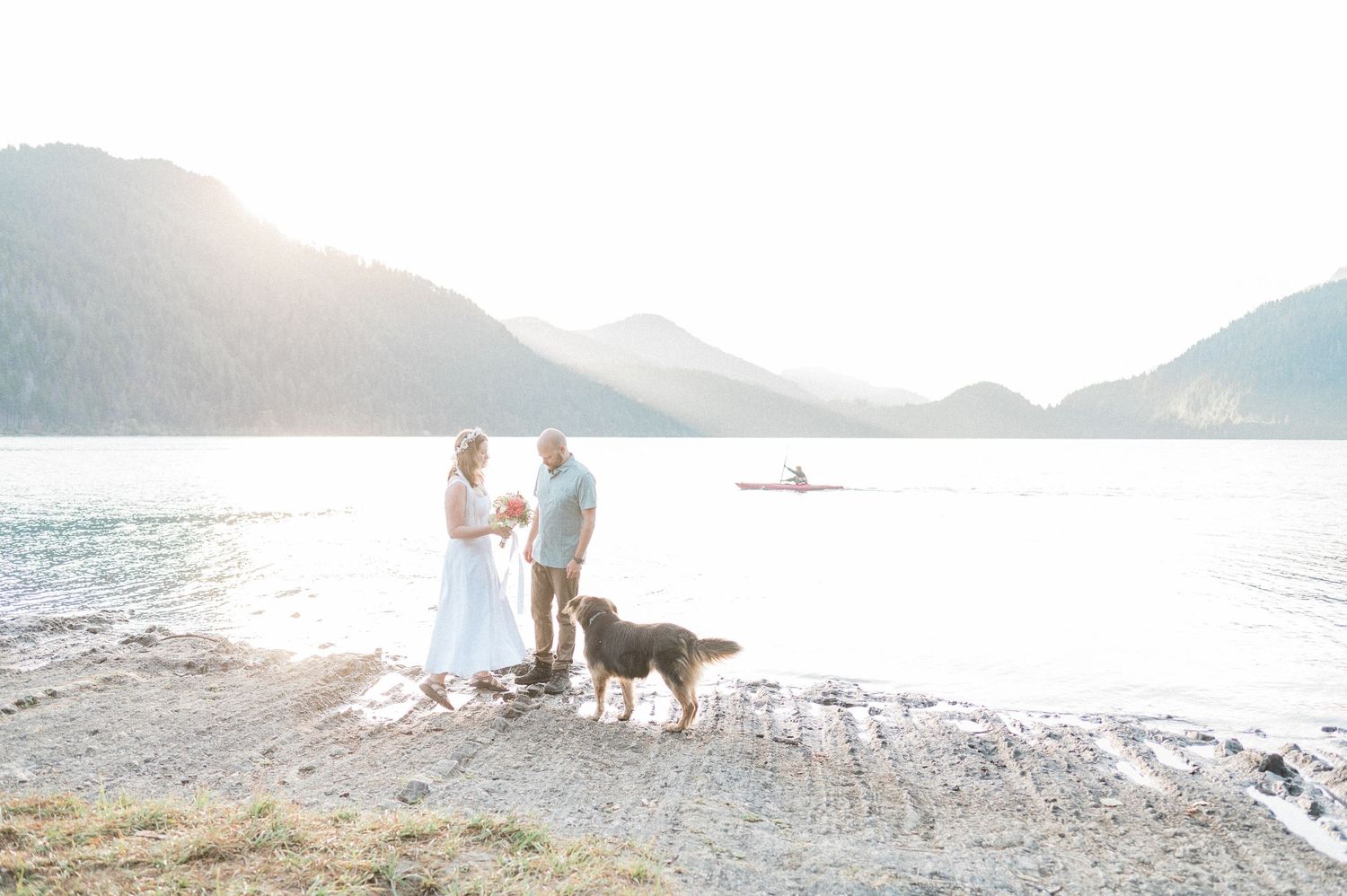 A bride and groom and their dog walk along a scenic beach at sunset with mountains in the background.