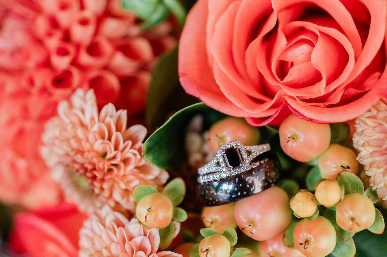 Close-up view of wedding rings among coral roses and pink dahlia flowers with delicate berries in a wedding bouquet arrangement.