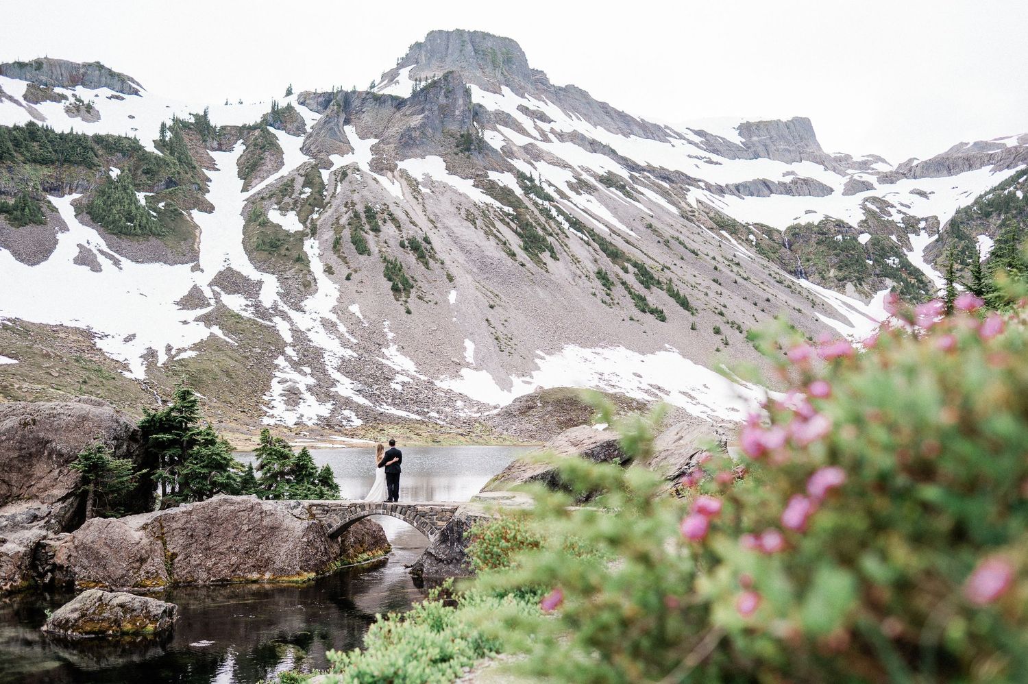 bride and groom stand on bridge at Heather Meadows with pink flowers in foreground and snowcapped mountain in background