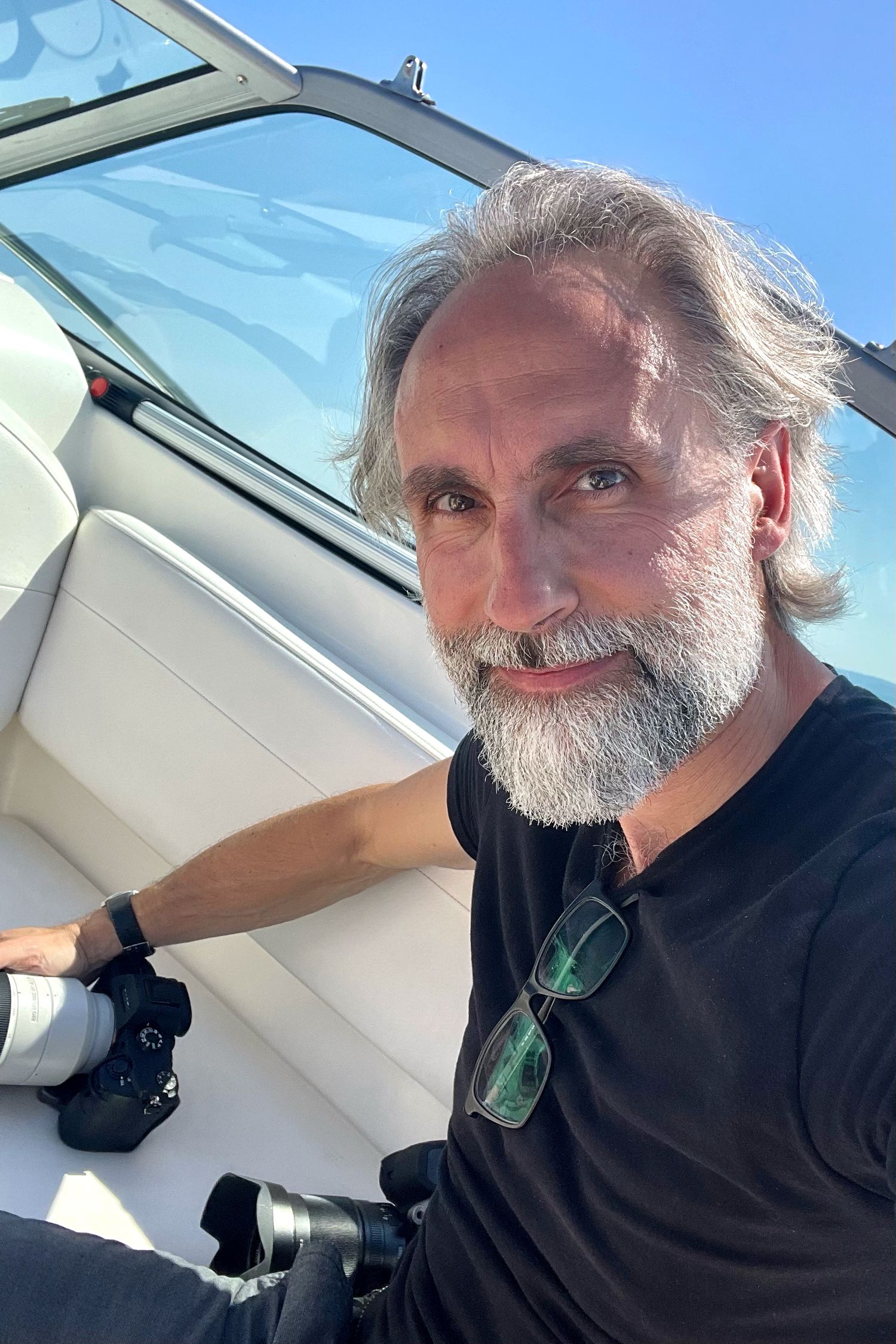 Bearded man in black shirt taking selfies inside a bright vehicle interior.