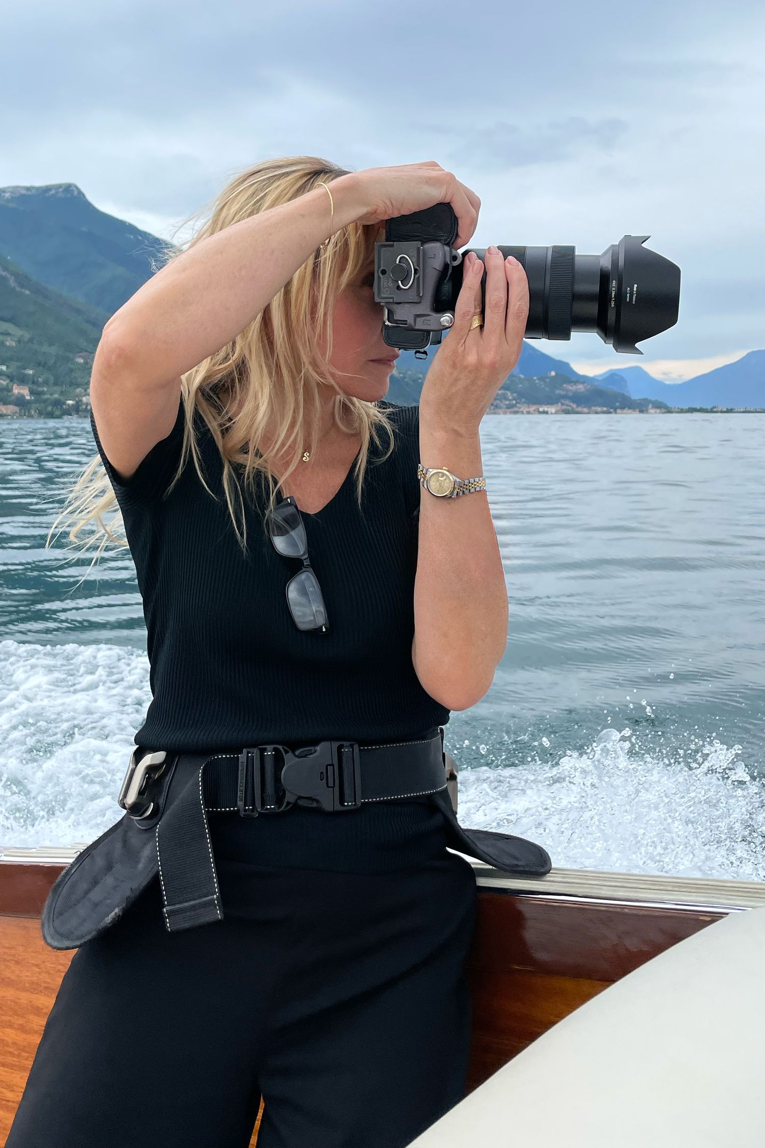 Photographer in black taking pictures from a boat with mountains and lake in background.