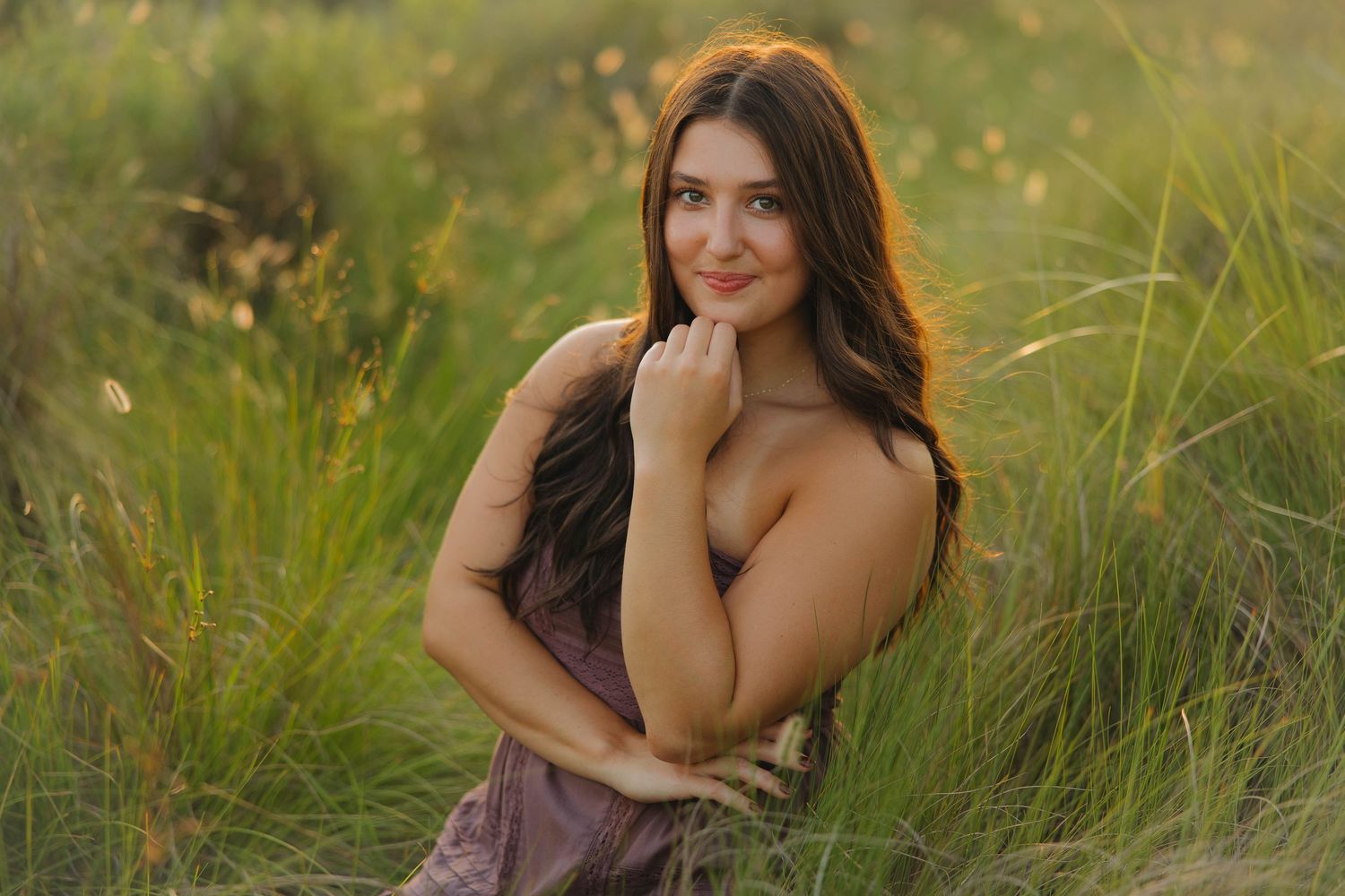 A smiling woman in a brown dress poses outdoors among tall grass during golden hour.