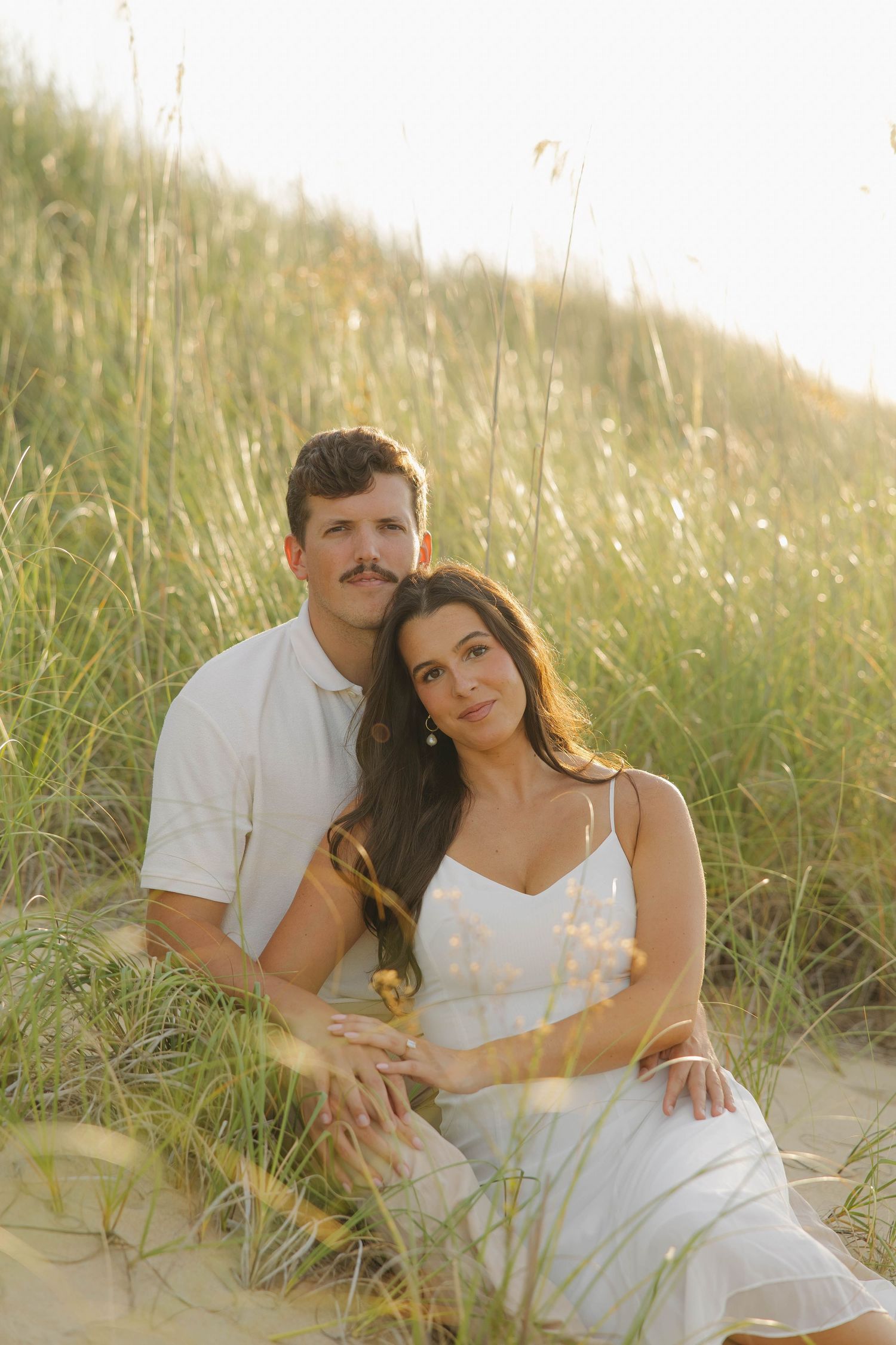 Romantic couple in white outfits sitting together on sandy beach at sunset in tall grass.