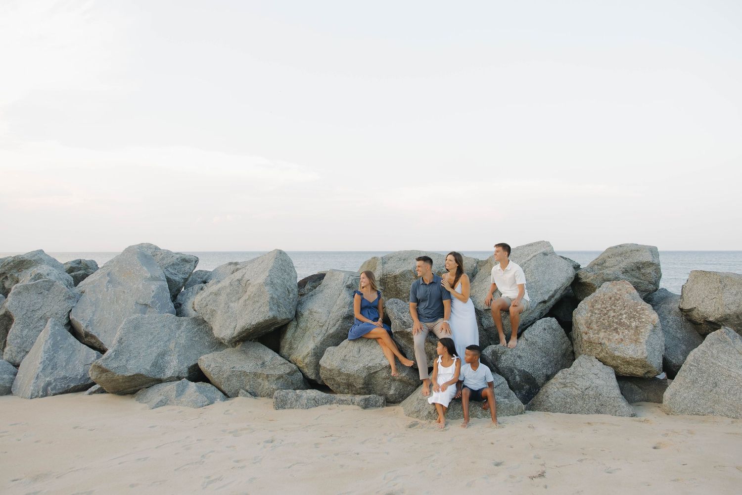 Family relaxing on large coastal rocks at sunset with ocean and beach view in the background.