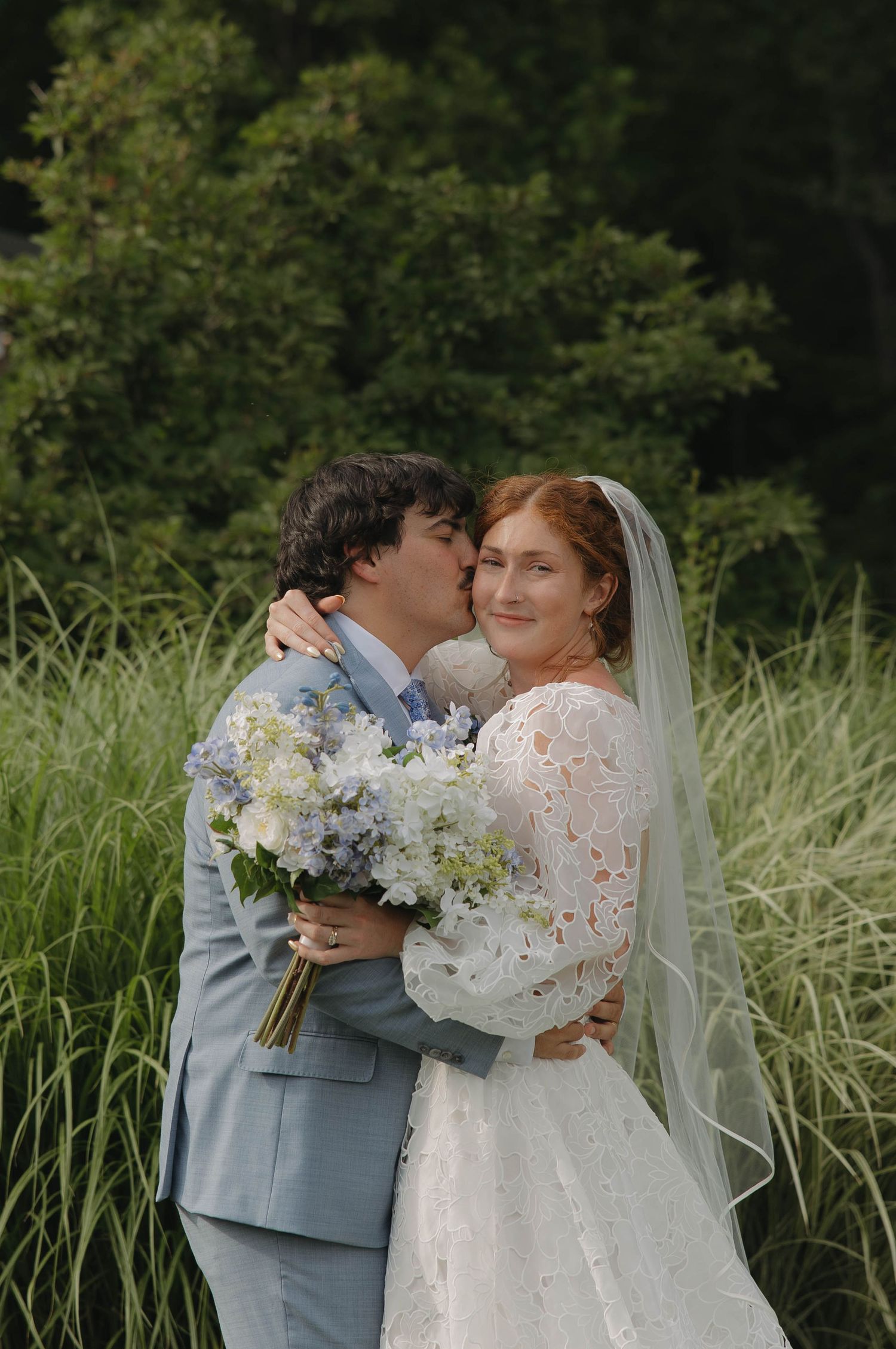 Romantic outdoor wedding scene with couple embracing in front of lush greenery and holding delicate white floral bouquet.