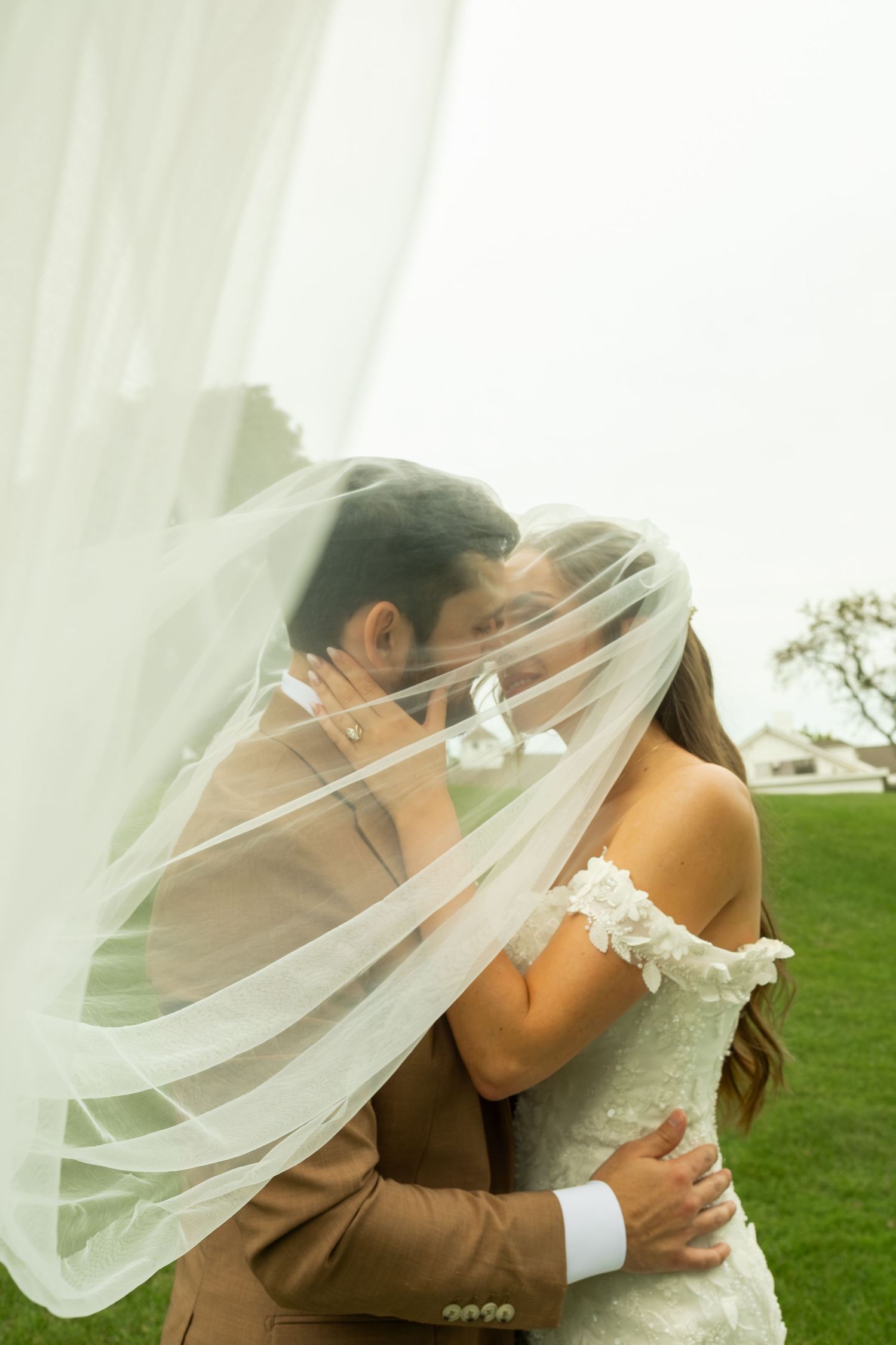 Bride and groom share a kiss beneath a flowing sheer wedding veil on a green lawn during an overcast outdoor wedding photo session.