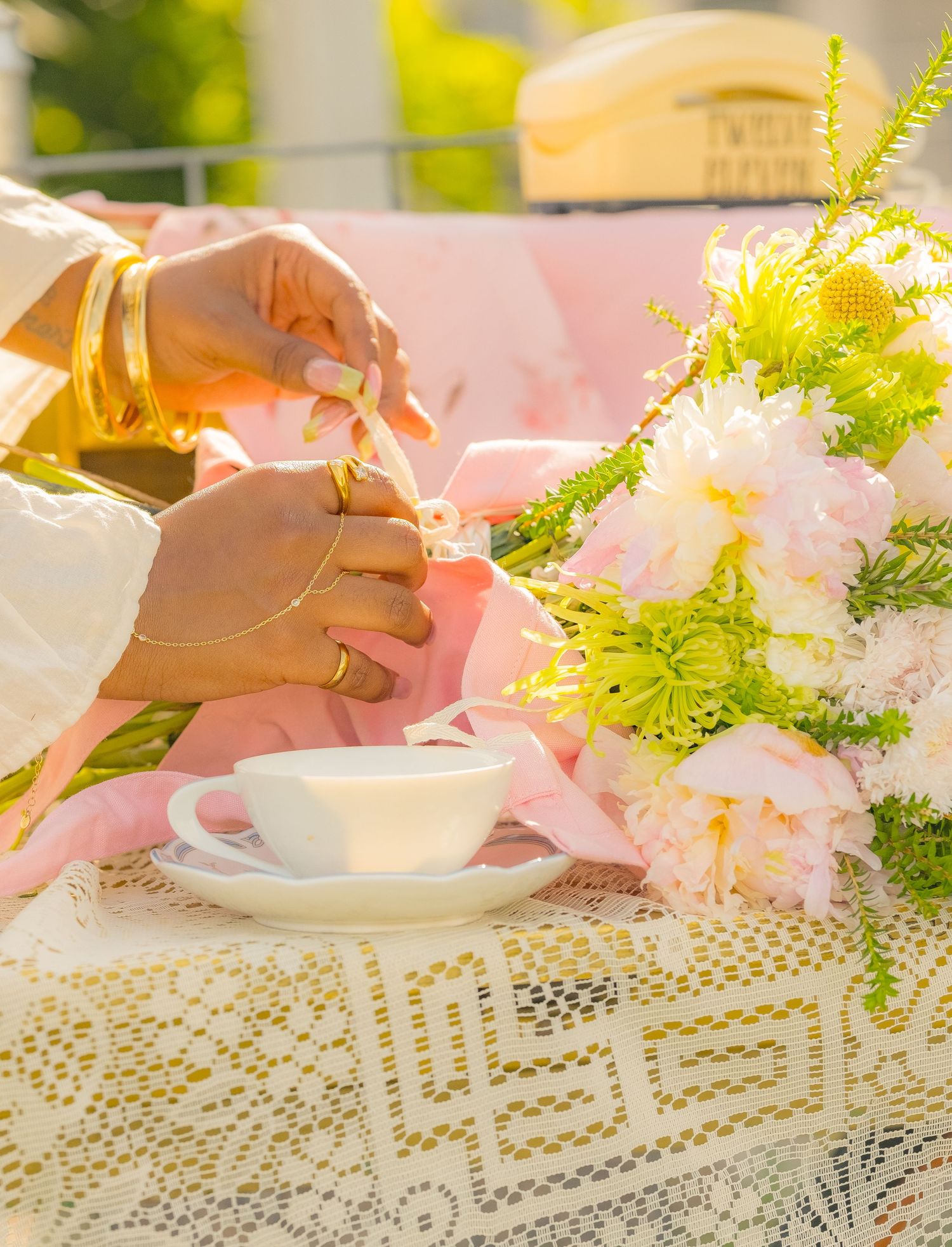 Elegant tea party setup with pink peonies, gold bangles, white teacup, and lace tablecloth in soft pastel tones.