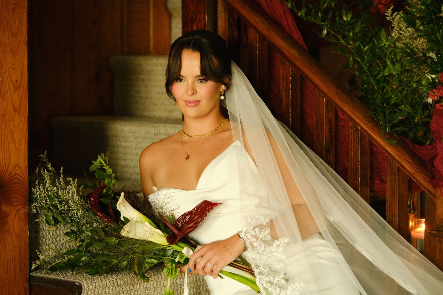 A bride in a white wedding dress poses on a rustic wooden staircase surrounded by greenery and candlelight.