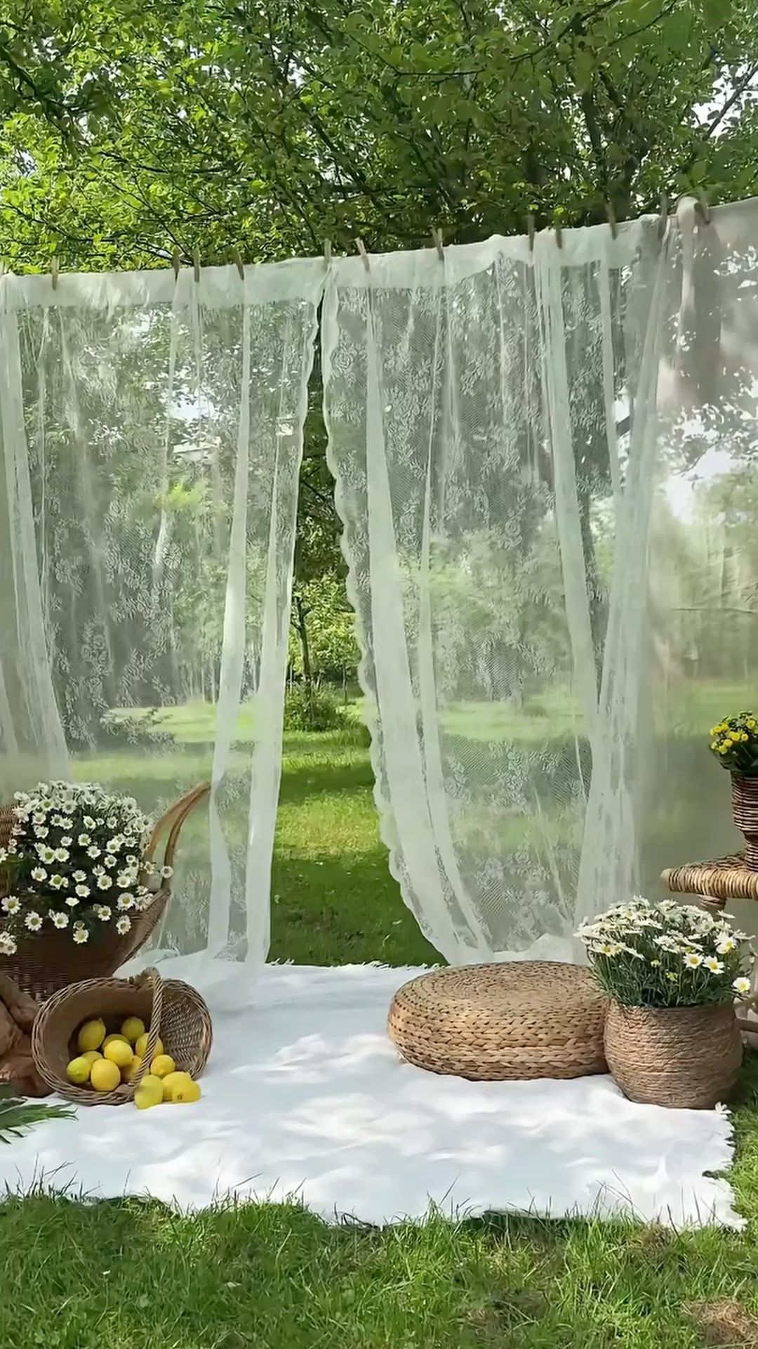 Sheer white curtains draped outdoors with daisies, lemons, and wicker baskets on a white blanket in a lush garden.