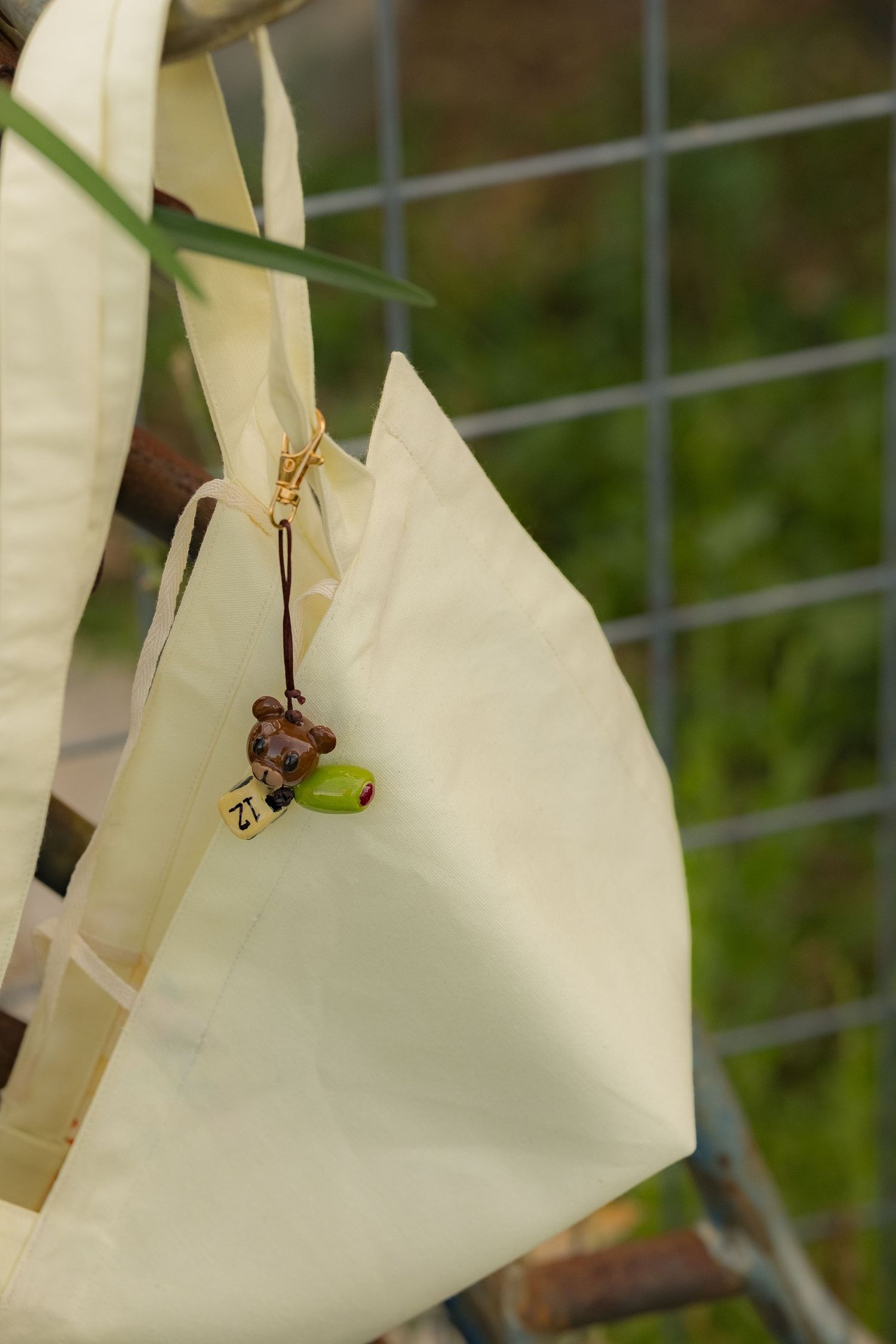 Close-up of a cream tote bag with a small acorn charm hanging from a wire fence outdoors.