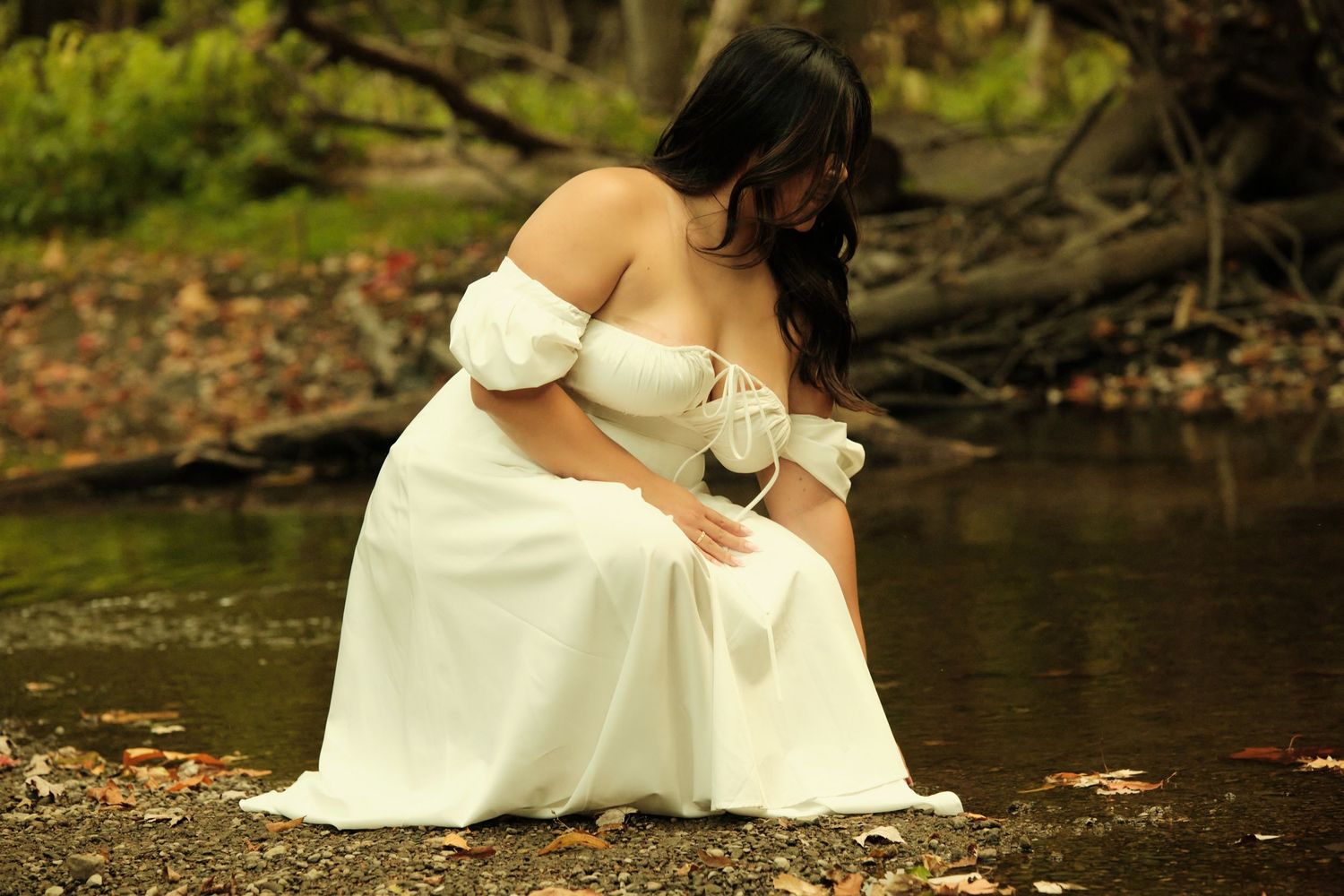 A bride in a white wedding dress sits gracefully on fallen autumn leaves near a tranquil pond.