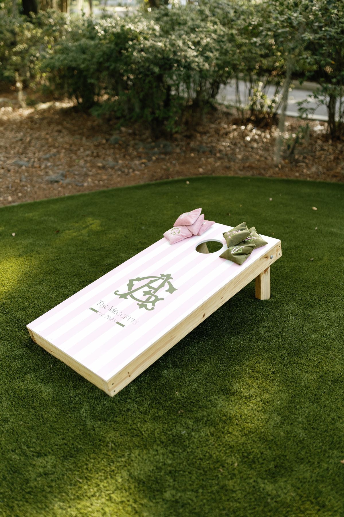 Custom pink striped cornhole board with monogram logo on green artificial turf, with pink and green bean bags.