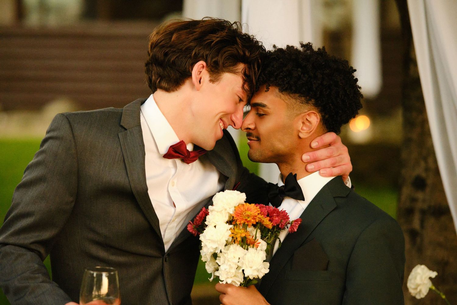 A couple in wedding attire share an intimate moment with white flowers and a rustic outdoor setting in the background.